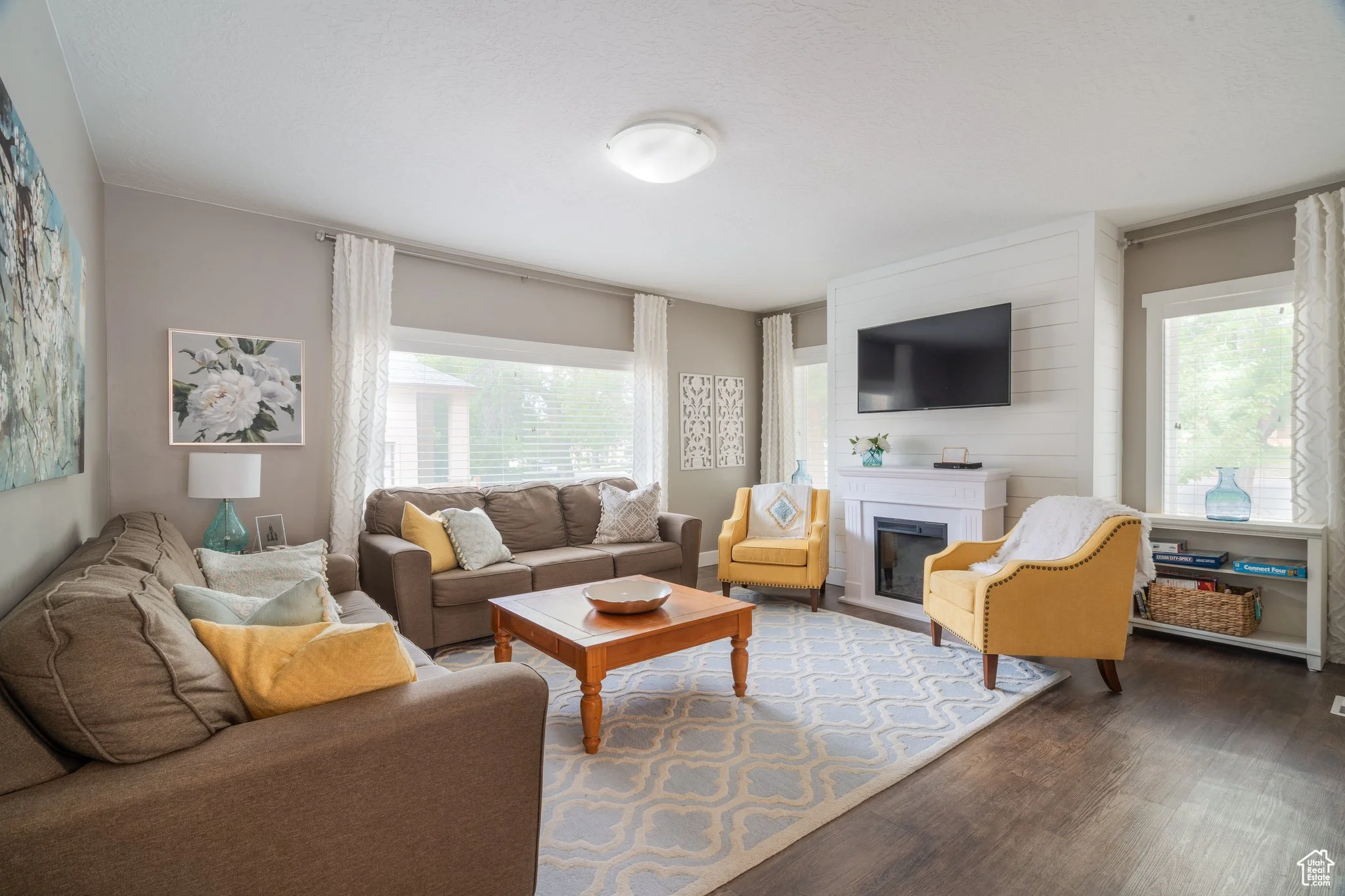 Living room with beige and yellow armchairs, a brown sofa with pastel pillows, a wooden coffee table, a patterned rug, a white fireplace with a TV above, windows with white curtains, and decorative vases on the window sills.