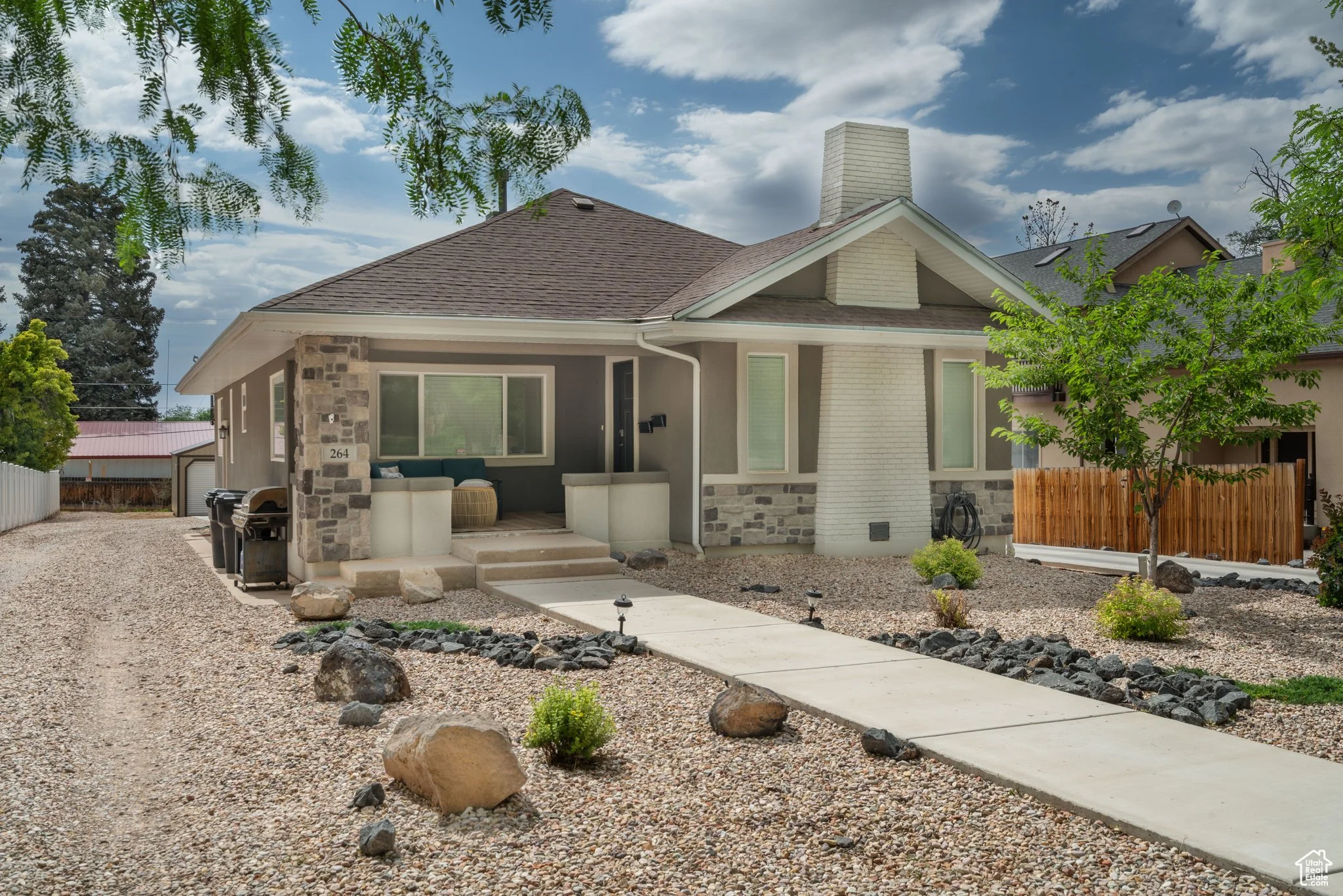 A modern single-story house with gray and white exterior, stone accents, large windows, a driveway on the left, a concrete pathway in front, a young tree, and a landscaped yard with rocks and small plants under a partly cloudy sky.
