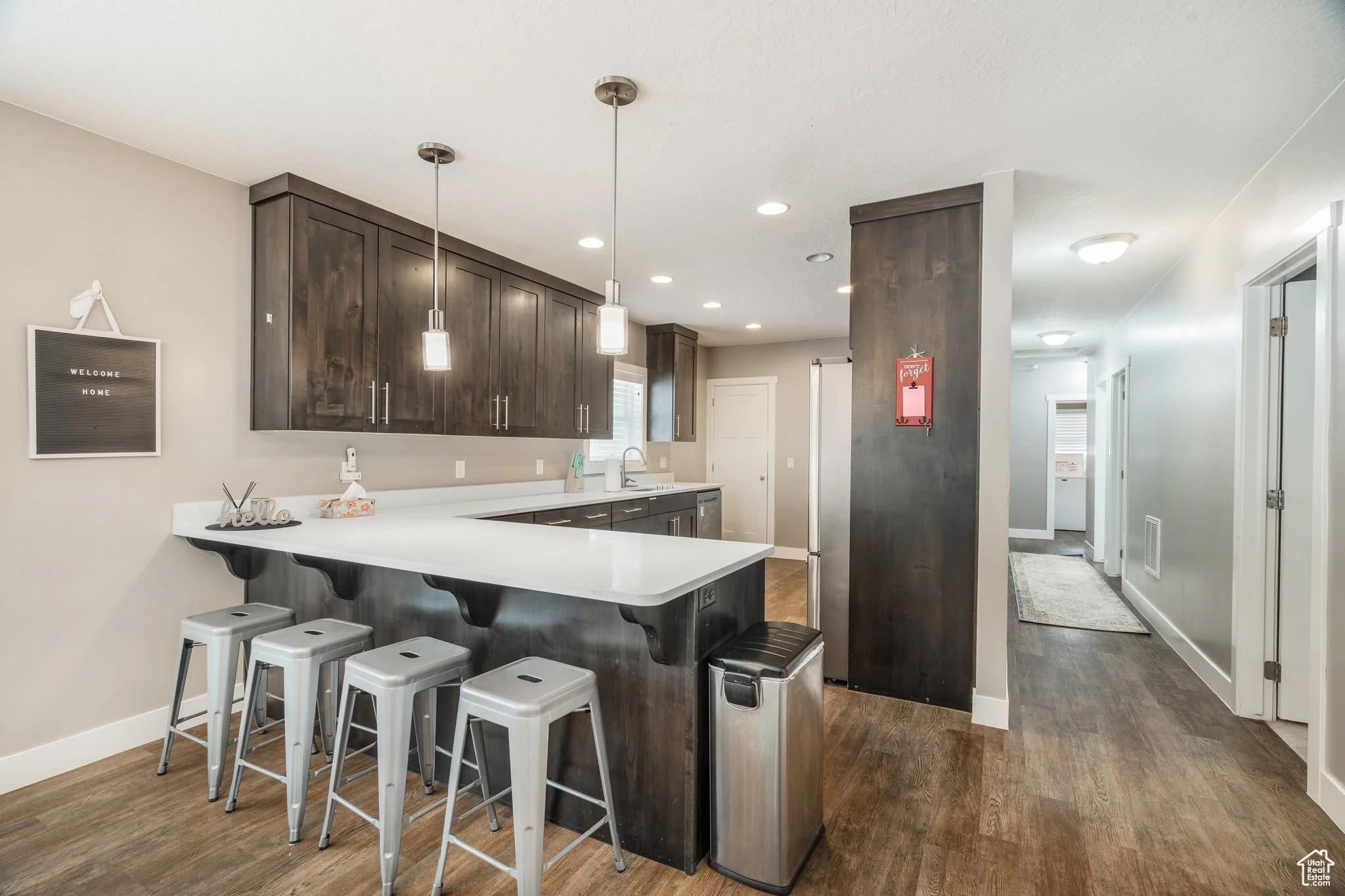 Modern kitchen with dark wood cabinets, white countertop, and four metal barstools at the island. Pendant lights hang above the island, and a window over the sink lets in natural light. A hallway leads to other rooms.
