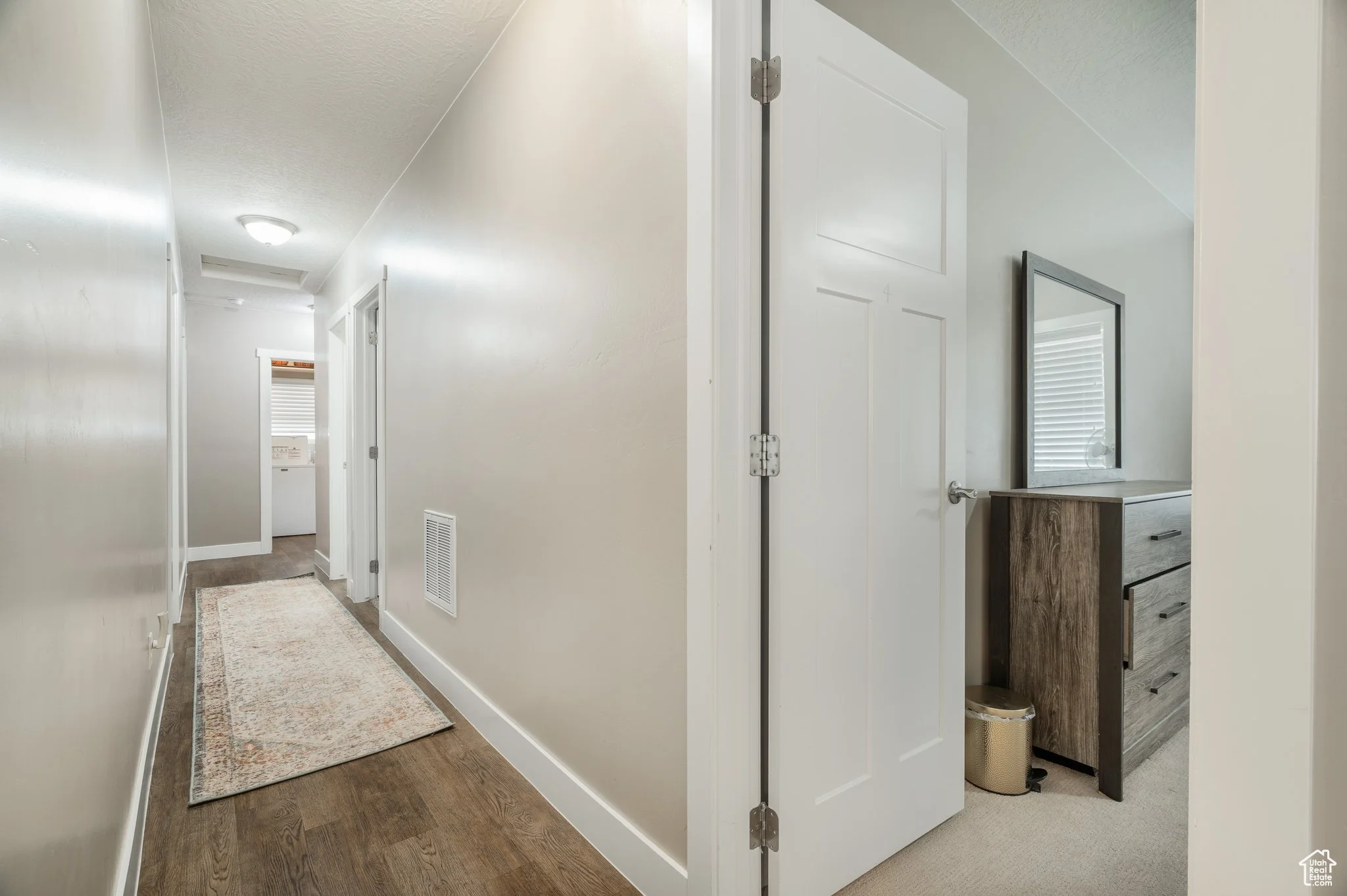 A hallway with white walls, brown wooden flooring, a beige patterned rug, and ceiling light. An open door on the right shows a small room with a dresser, mirror, and a window with blinds. Further down the hall, there are baseboard vents and doorways,