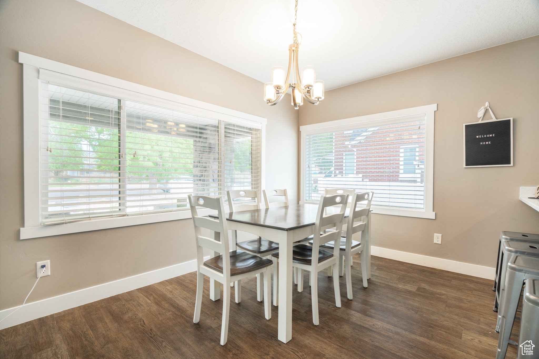 Bright dining room with a rectangular table surrounded by six white chairs with wood seats, beige walls, two large windows with white blinds, a hanging chandelier, a chalkboard sign on the wall that reads "Welcome Home," and part of a stainless steel