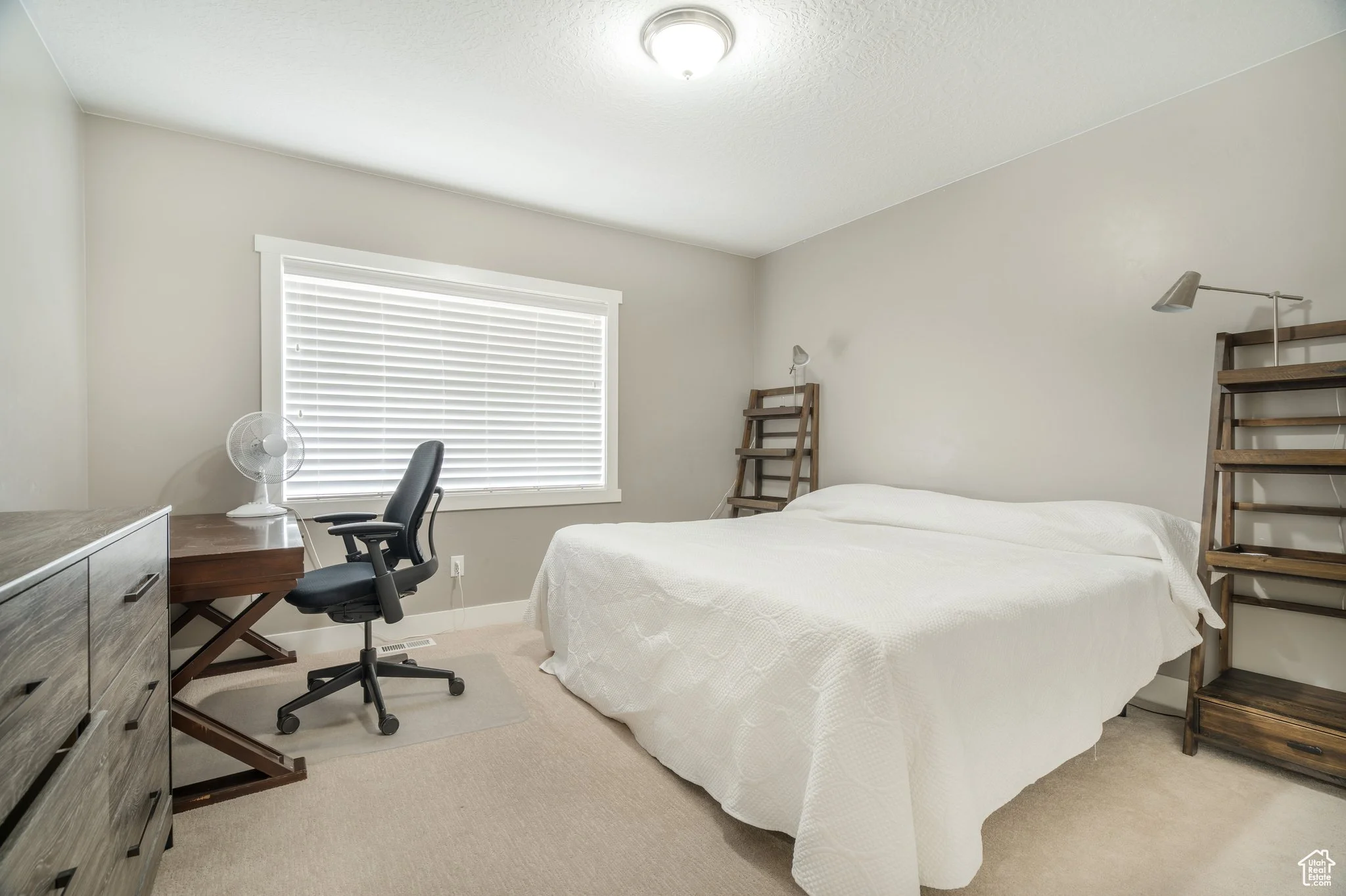 A bedroom with a bed covered in a white quilt, two wooden ladder-style shelves on either side of the bed, a window with closed white blinds, a black office chair next to a wooden desk, a small fan on the desk, a dresser on the left side of the room, and a beige carpeted floor.