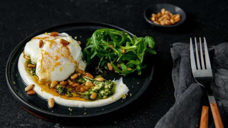 Poached eggs with spinach, pine nuts, and sauce on a black plate, garnished with arugula and served with a side of pine nuts.