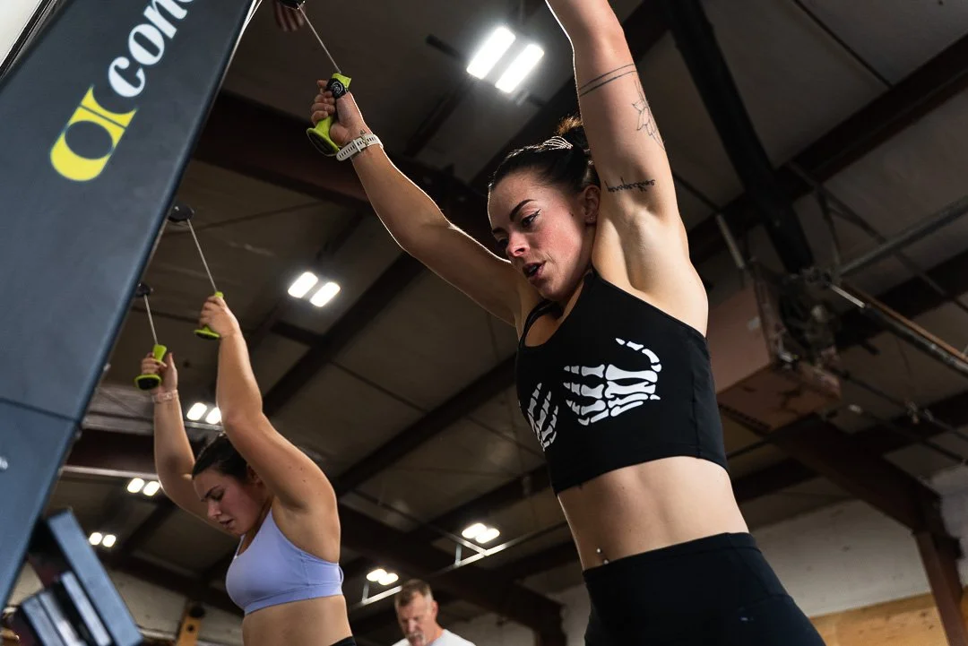 Two women working out at a gym, holding jump ropes overhead during an exercise. Both are wearing sports bras, and the gym has a high ceiling with exposed beams.