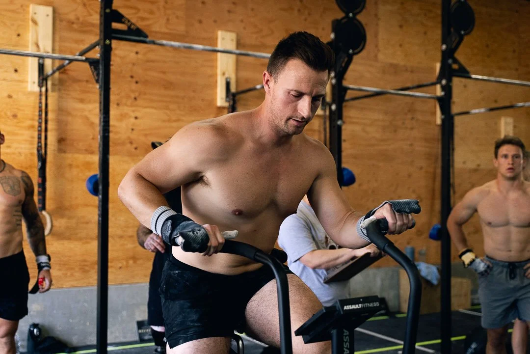 A shirtless man in black shorts using a stationary bike inside a gym, with other people in the background.