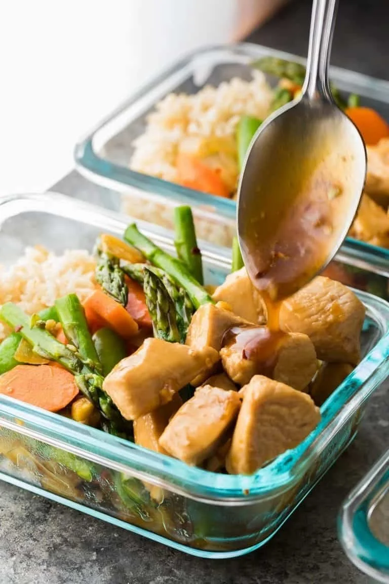 A glass container of cooked chicken pieces with gravy, accompanied by a vegetable medley of asparagus, carrots, and zucchini. In the background, another container with rice and vegetables is visible.