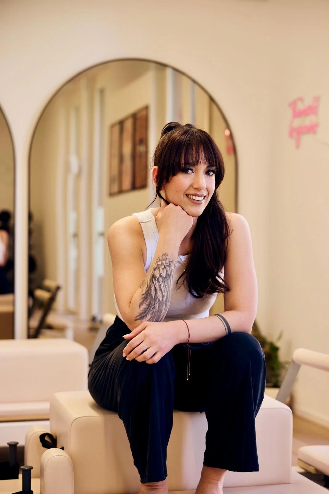 Junge Frau mit dunklen Haaren, Tattoo am Arm, sitzt auf einem weißen Sitzmöbel in einem Café, smiling und schaut in die Kamera.