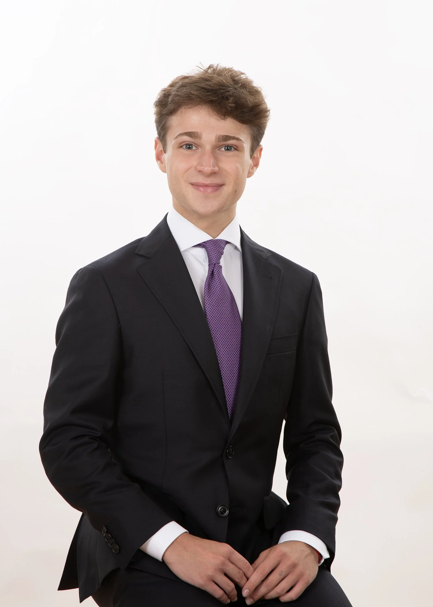 A young man with light brown hair and blue eyes, dressed in a black suit, white shirt, and purple tie, posing against a plain white background.