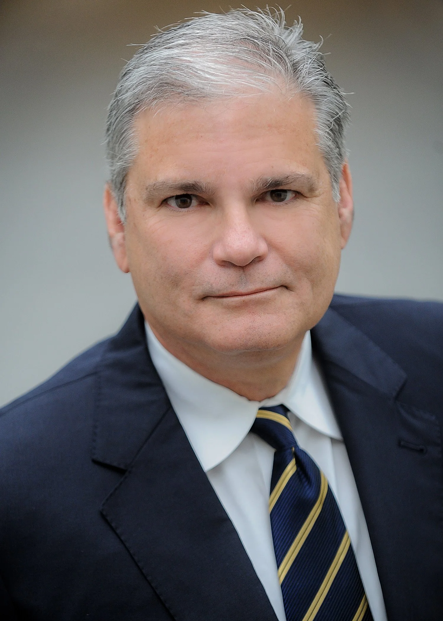 A middle-aged man with gray hair wearing a dark suit, white shirt, and striped tie.