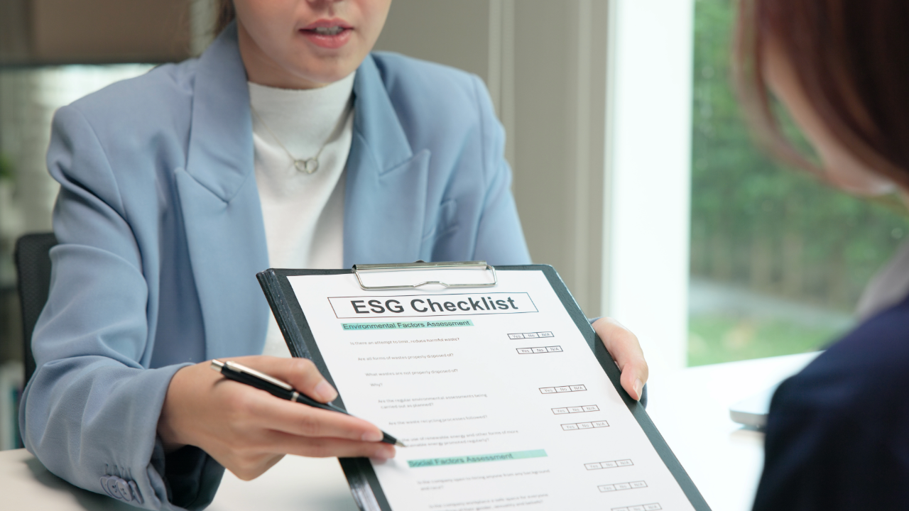 A woman in a light blue blazer showing an ESG Checklist on a clipboard to another person during a meeting.