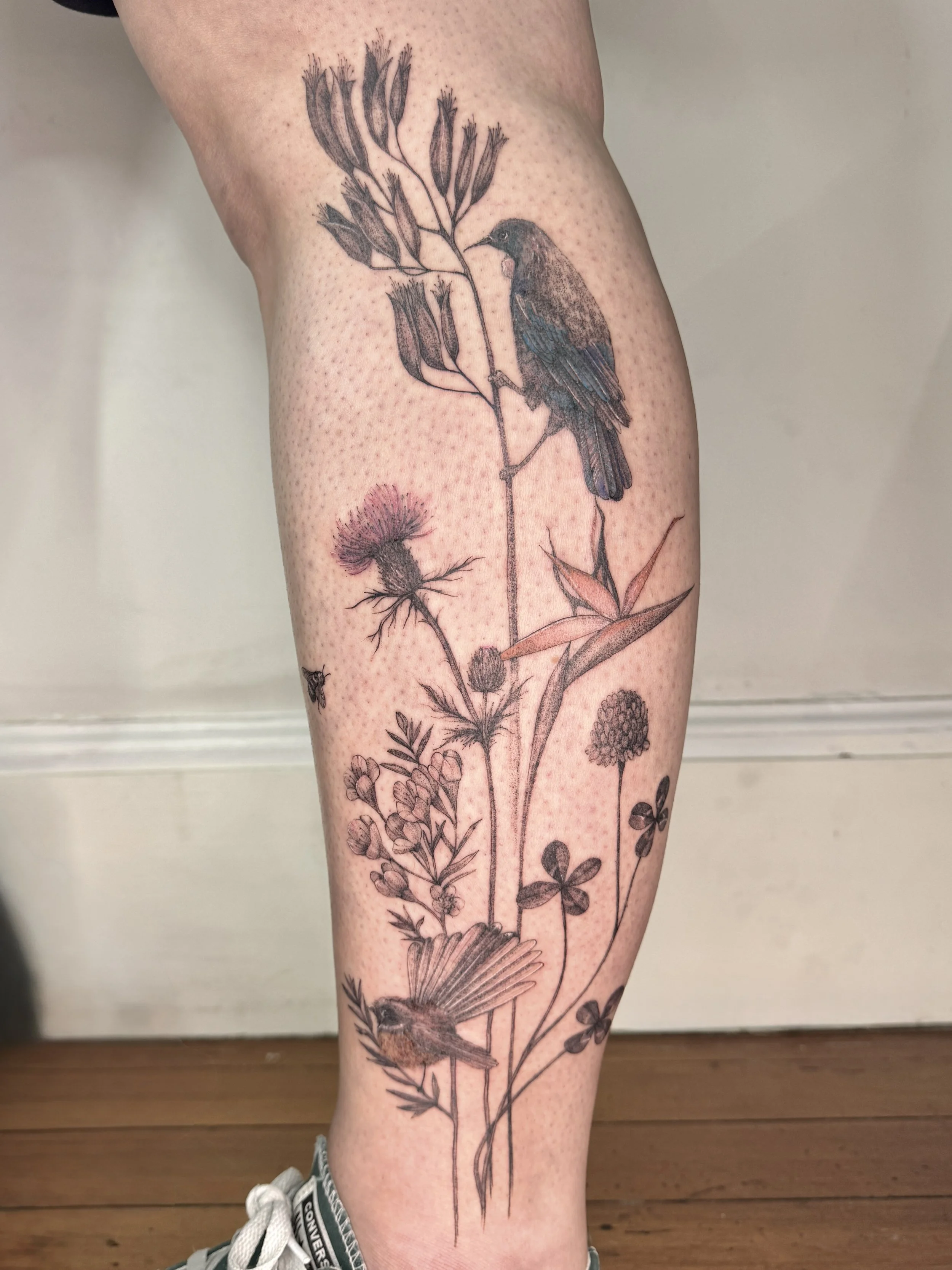 Tattoo of a bird perched on a branch among various native flowers and plants on a person's leg