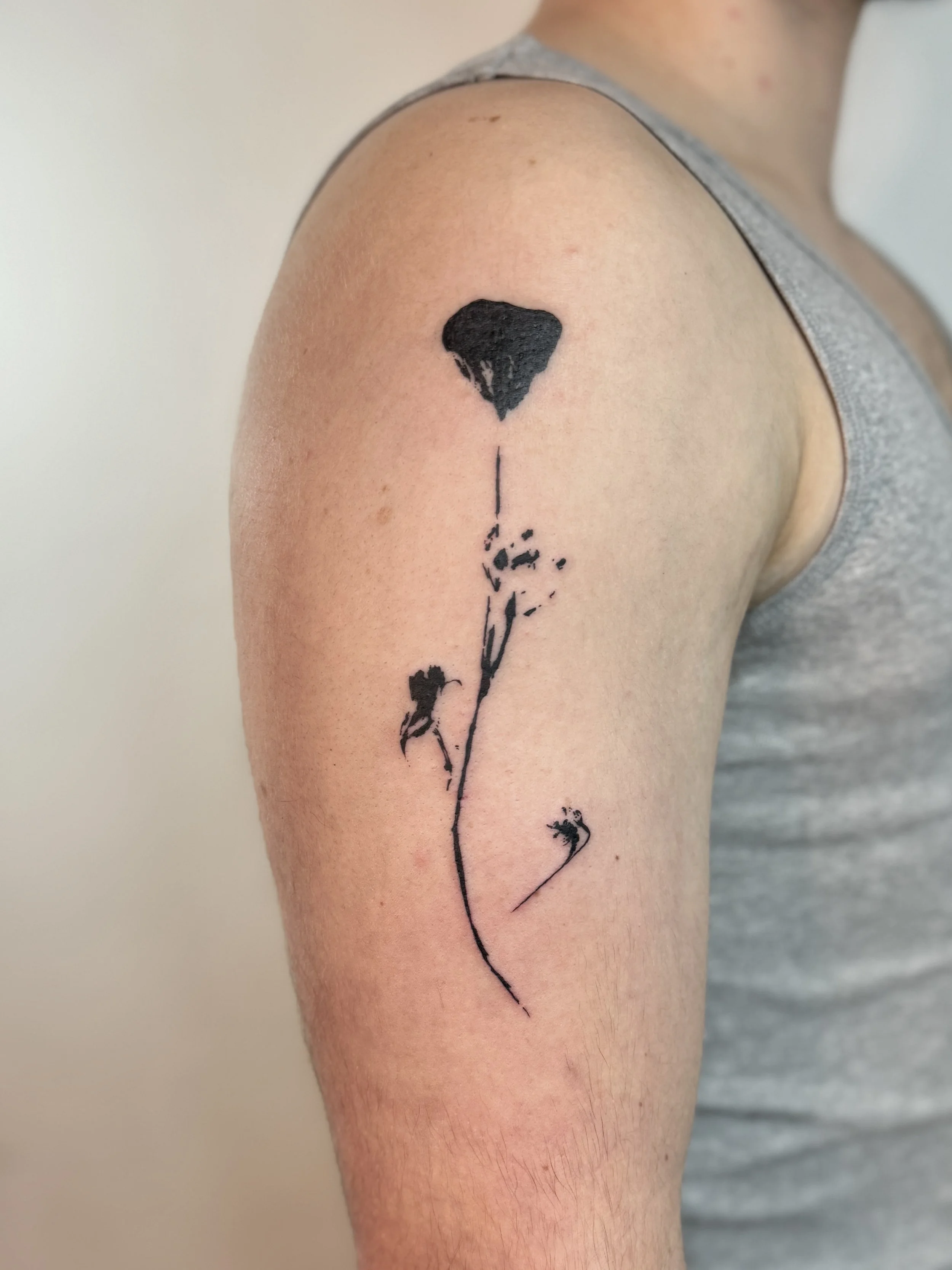 Black ink tattoo of a Californian poppy with a long, curved stem on a person's upper arm. Close-up of botanical tattoo texture and linework, fine line tattoo Vancouver
