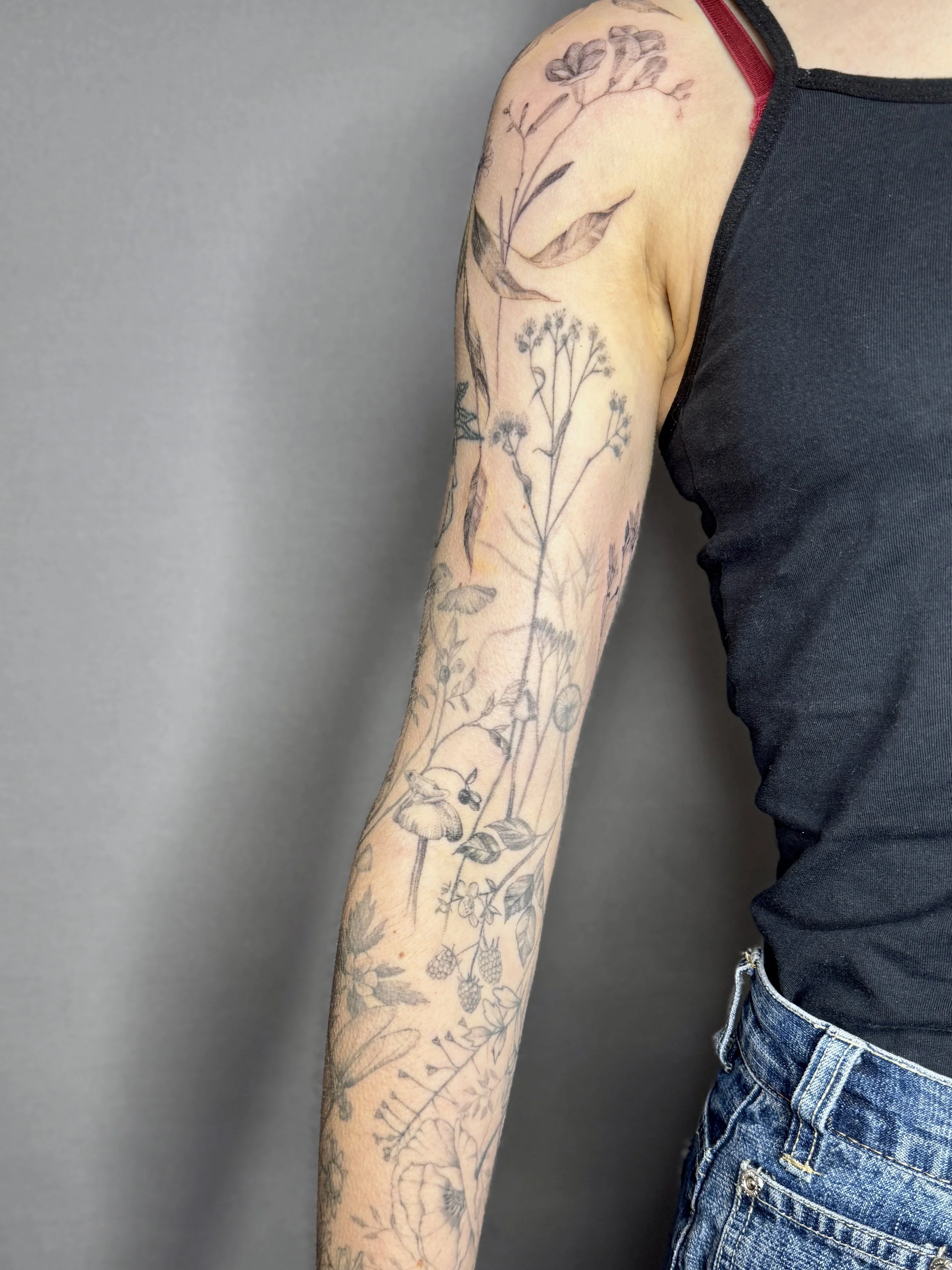 A person's upper arm and shoulder with a detailed black and gray fineline floral tattoo. Fine line botanical flash tattoo design, tattoo artist in Vancouver