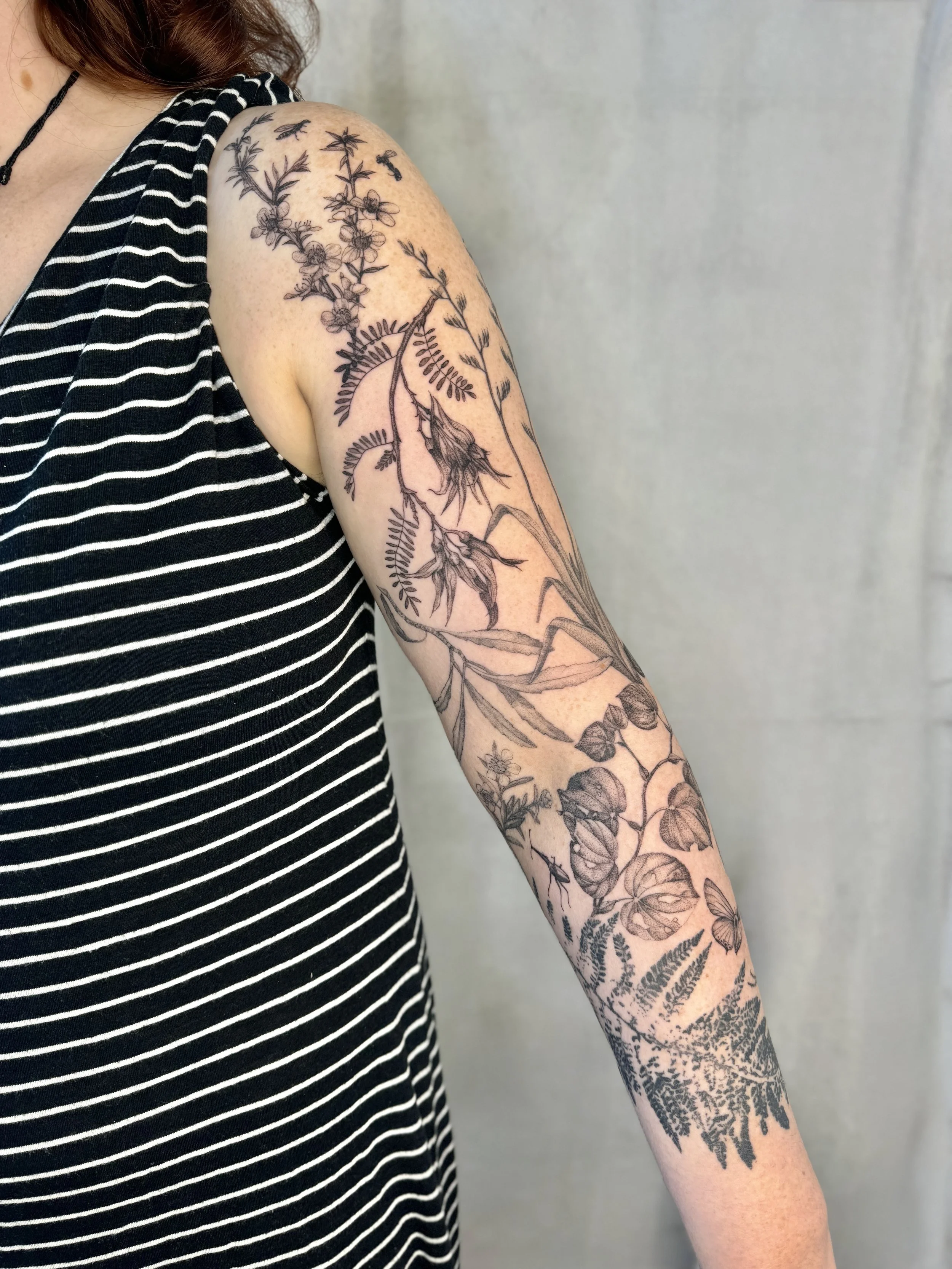 A person's upper arm and shoulder with a detailed black ink floral tattoo, including various flowers, leaves, and a butterfly.Custom botanical sleeve, Vancouver tattoo artist