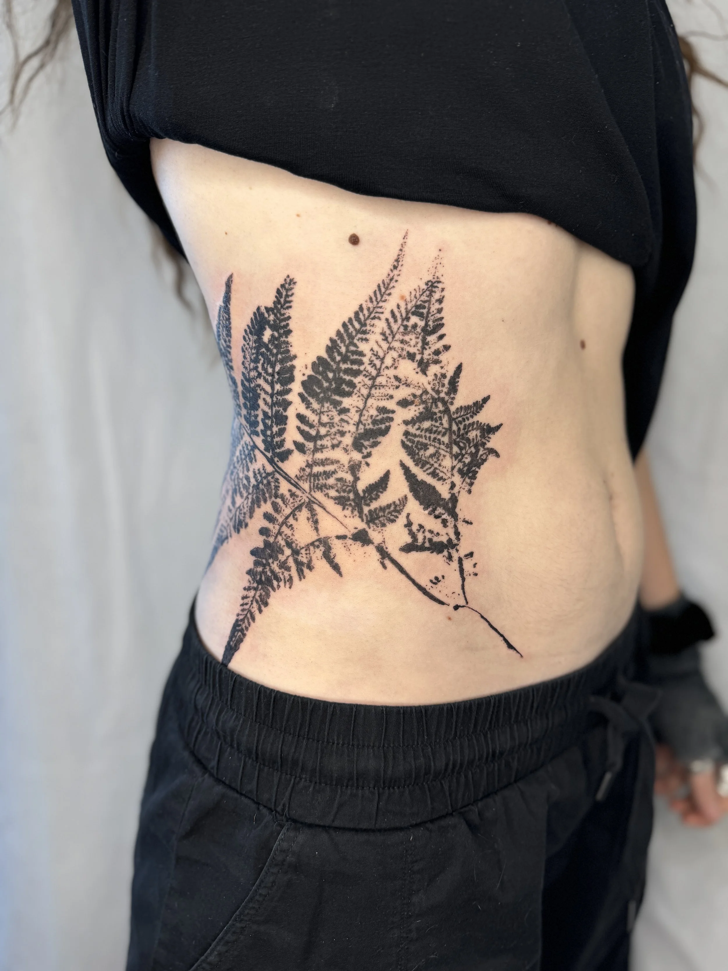 A person's side torso featuring a large black fern leaf tattoo. leaf imprint tattoo with minimalist composition, tattoo artist Vancouver