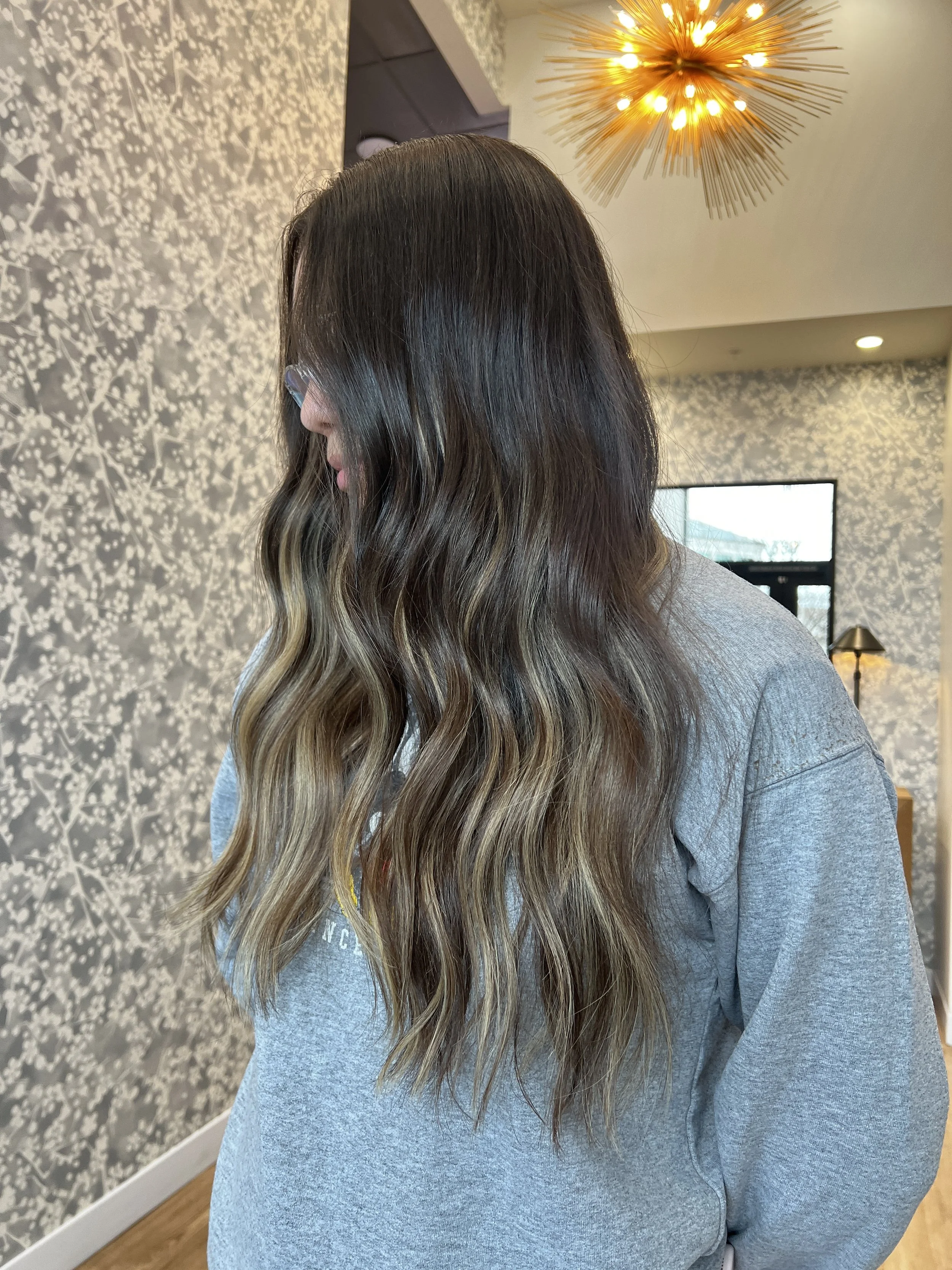 A woman with long, wavy, dark blonde hair with highlights, wearing glasses and a gray sweatshirt, standing indoors in a room with floral wallpaper and contemporary lighting.