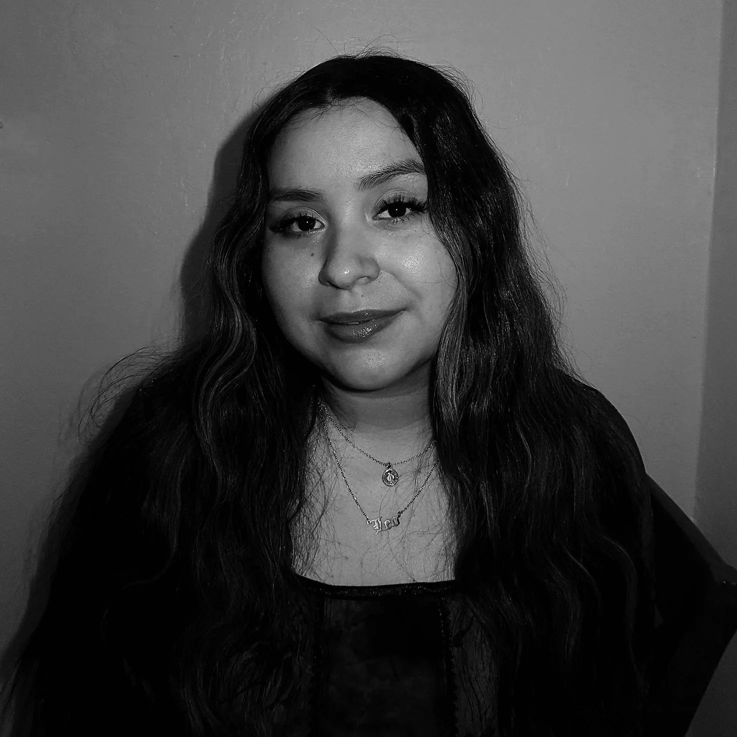 Black and white photo of a woman with long wavy hair, wearing a dark top and layered necklaces, smiling slightly.