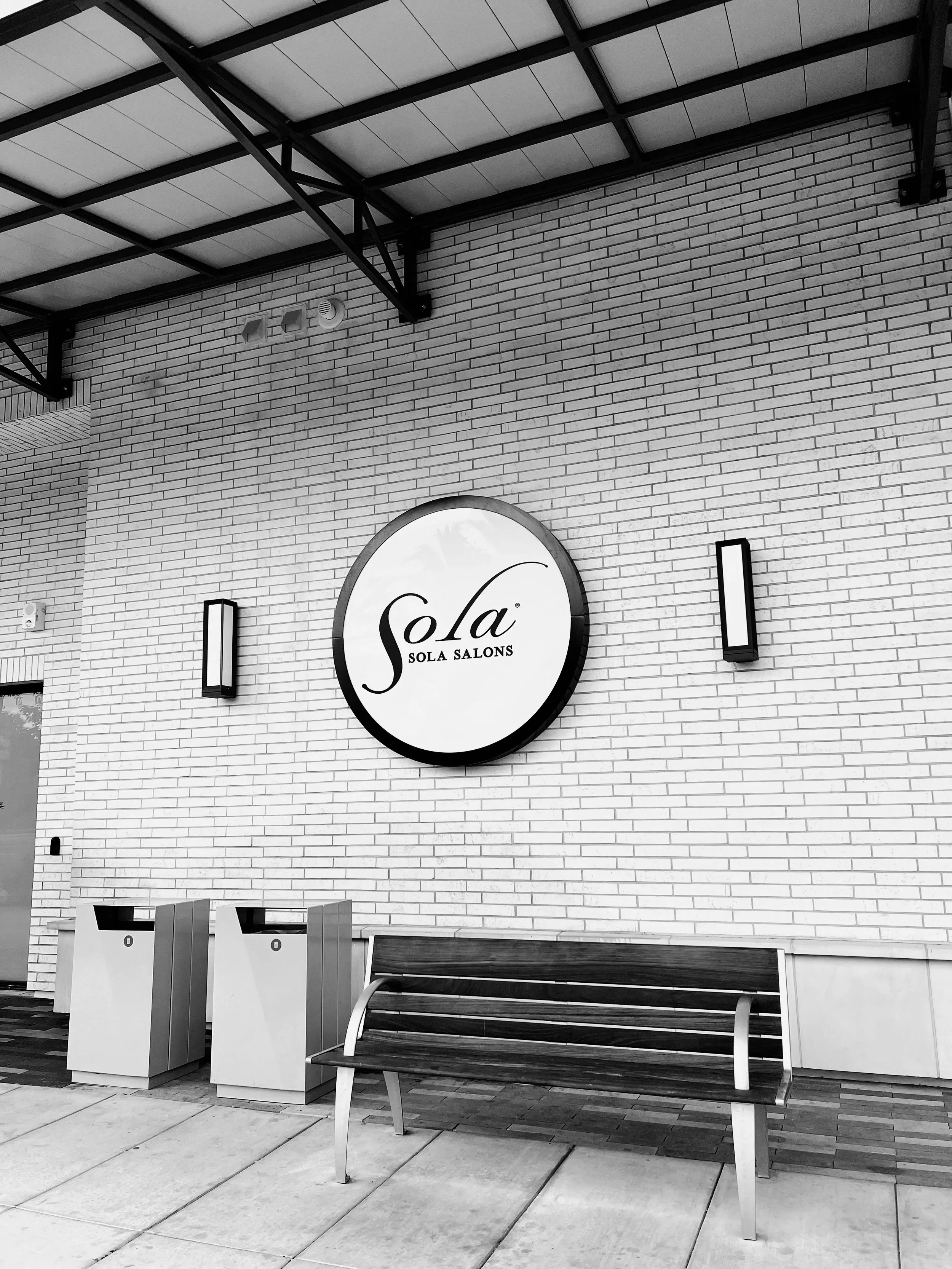 Exterior view of a salon with a circular sign reading 'Sola Salons,' a wooden bench, two trash bins, and wall-mounted lights on a brick wall.