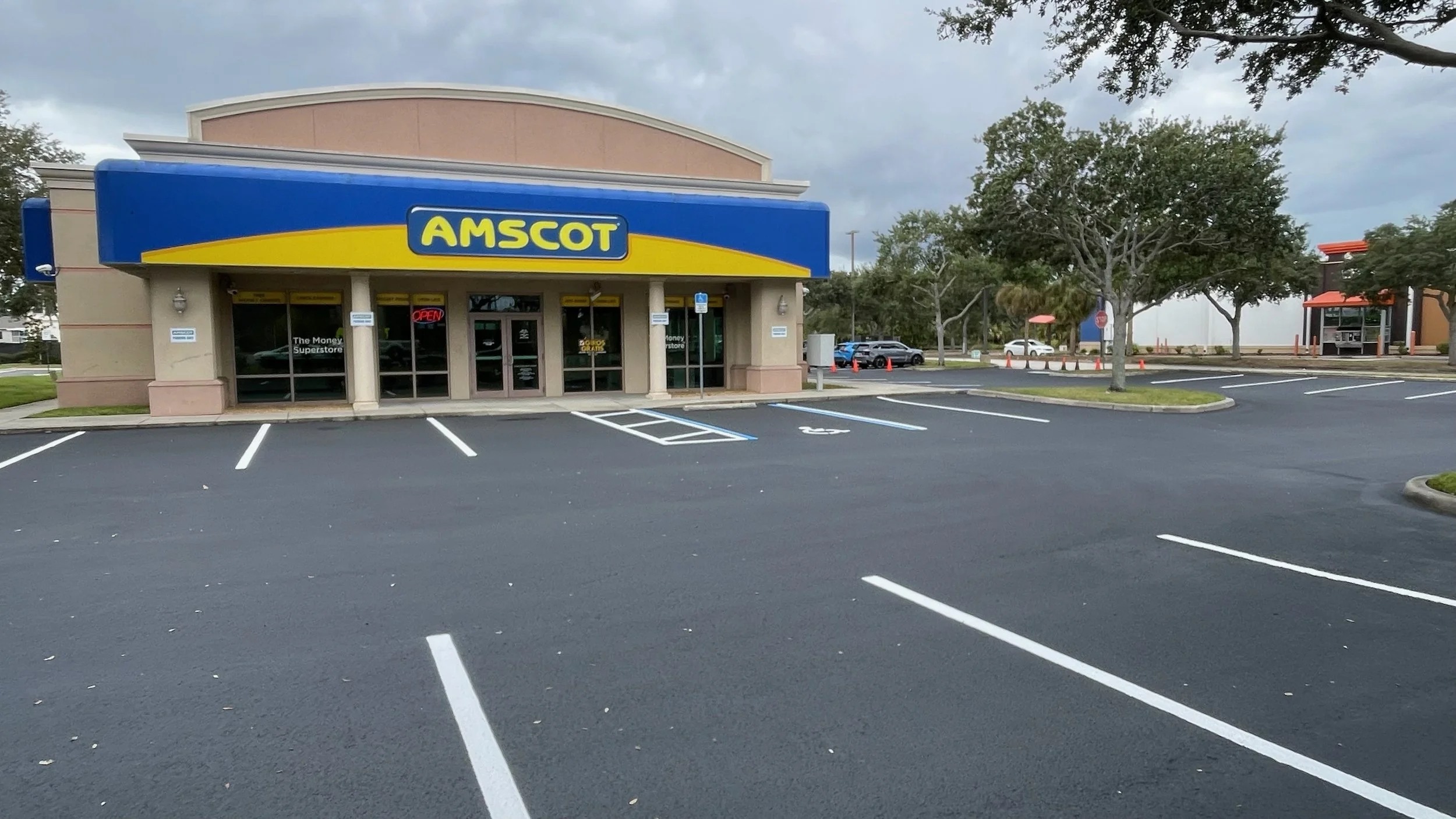 Freshly redone parking lot in front of an AMSCOT storefront with a blue and yellow sign, surrounded by trees and a cloudy sky.