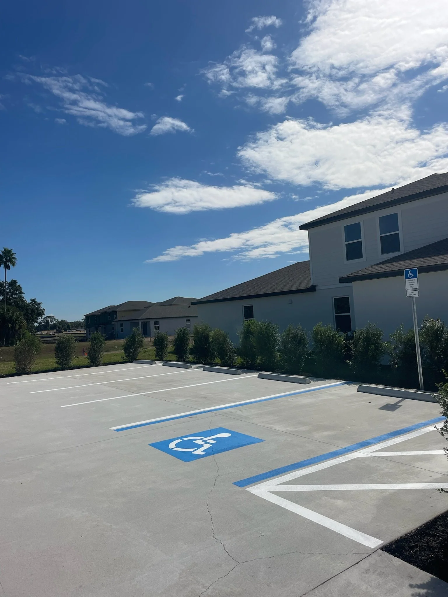 Freshly re-striped parking lot with designated handicapped parking space and sign, residential houses in the background, under a partly cloudy sky.