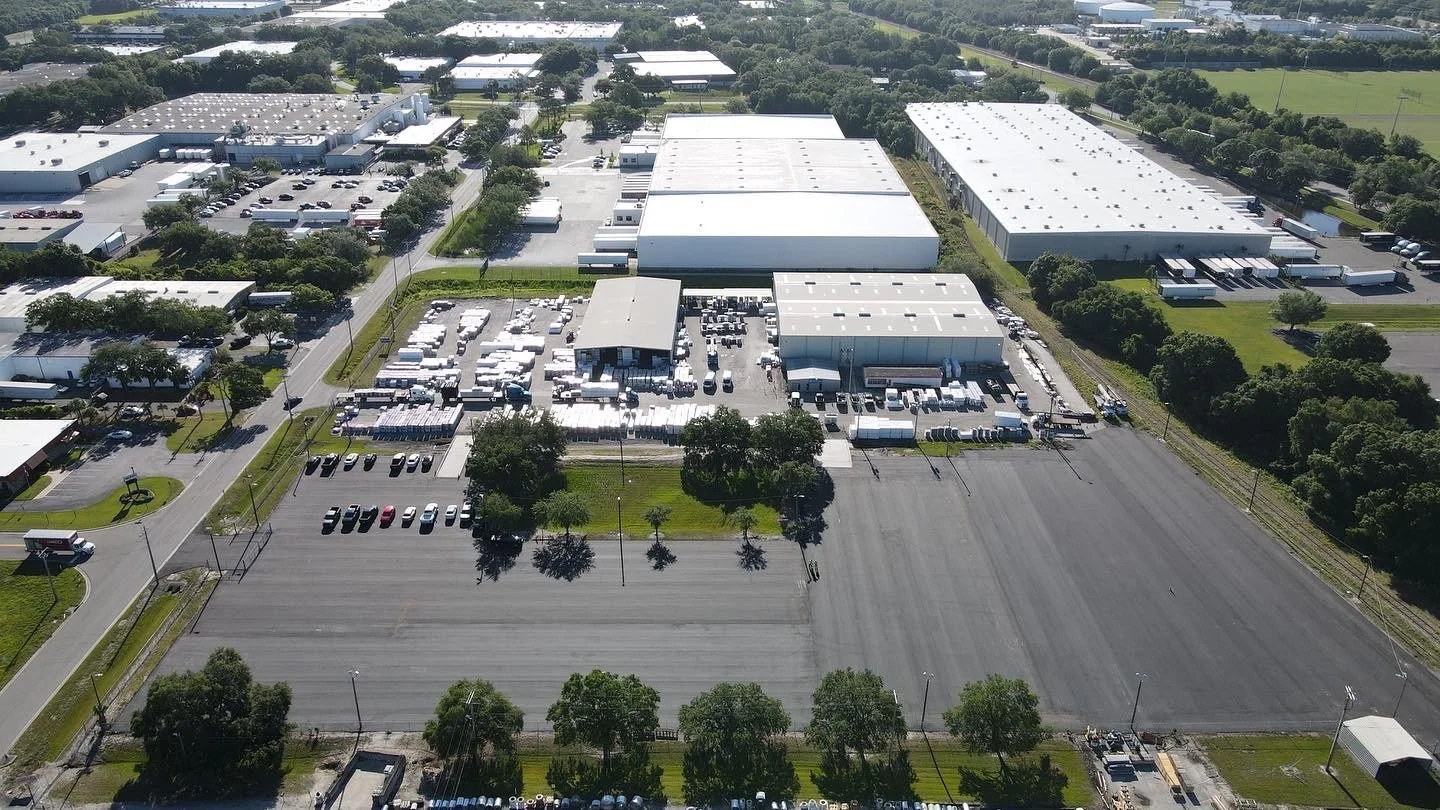 Aerial view of an industrial warehouse complex with multiple large buildings, parking lots, and outdoor storage areas, surrounded by green spaces and trees.