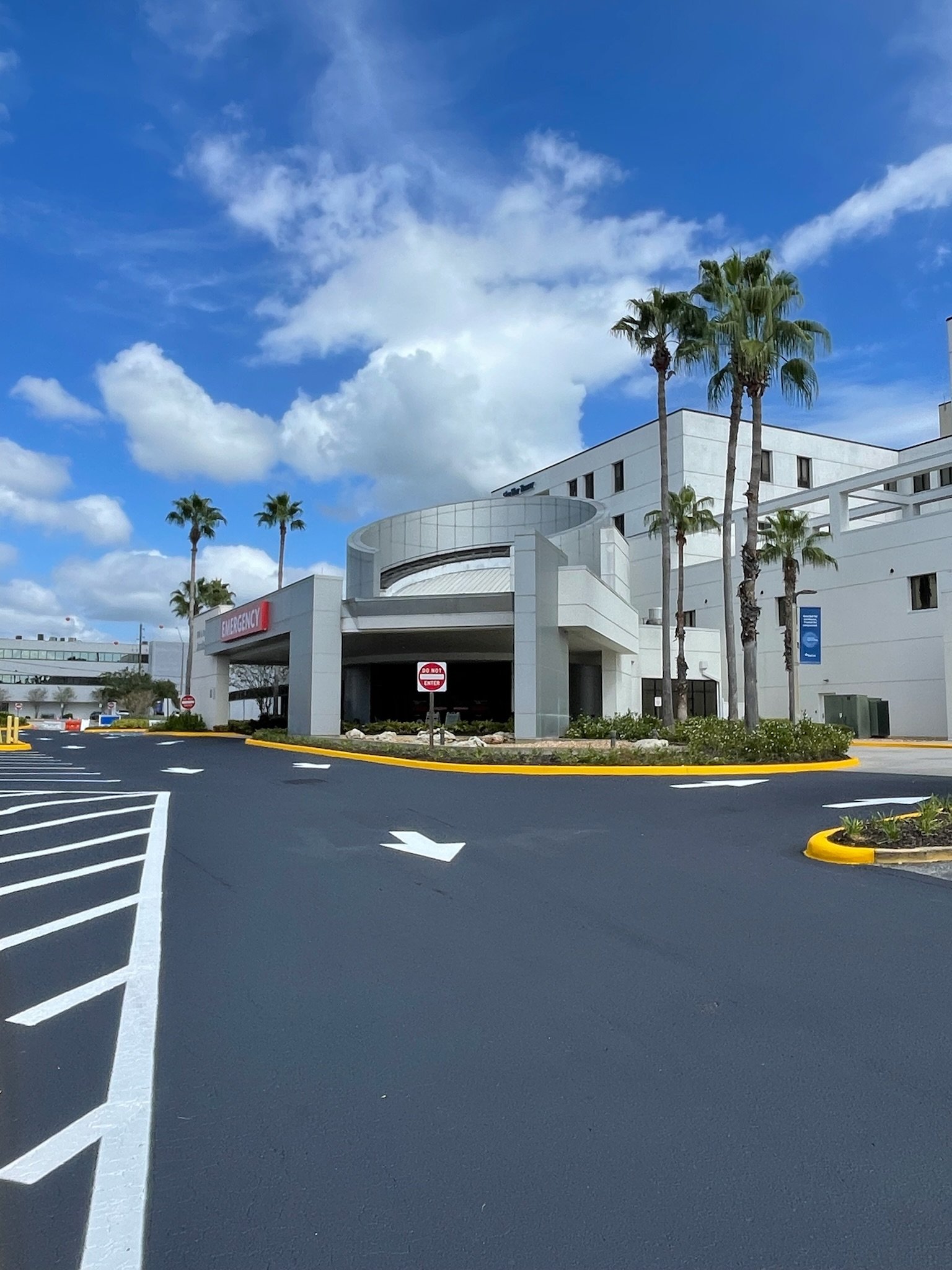Exterior view of a modern hospital emergency department with a white facade, palm trees, clear blue sky with clouds, and parking lot with directional arrows.