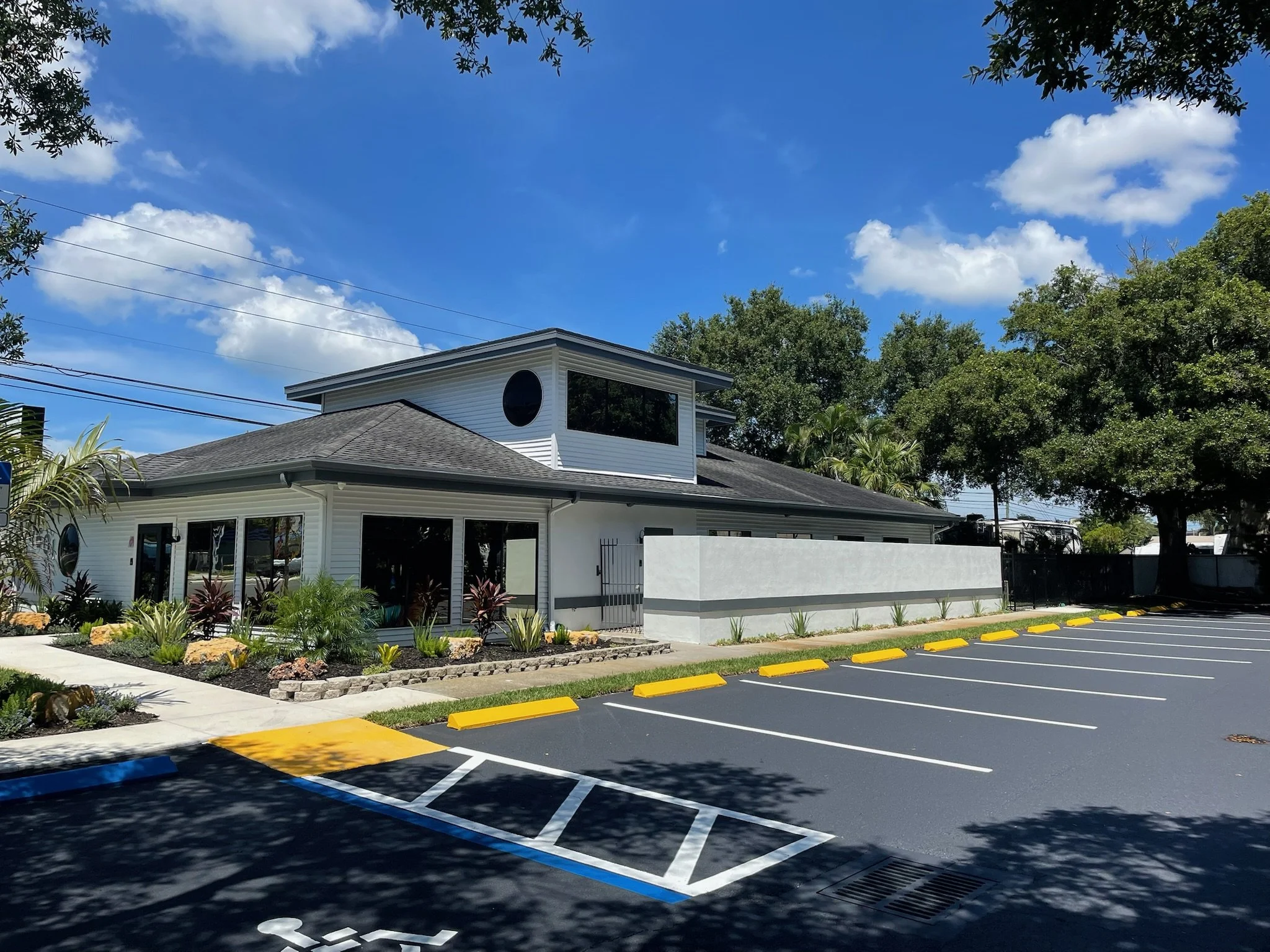 A modern building with white siding and a gray roof, located next to a parking lot with freshly painted yellow and blue curbs and parking lines, surrounded by trees and a partly cloudy blue sky.