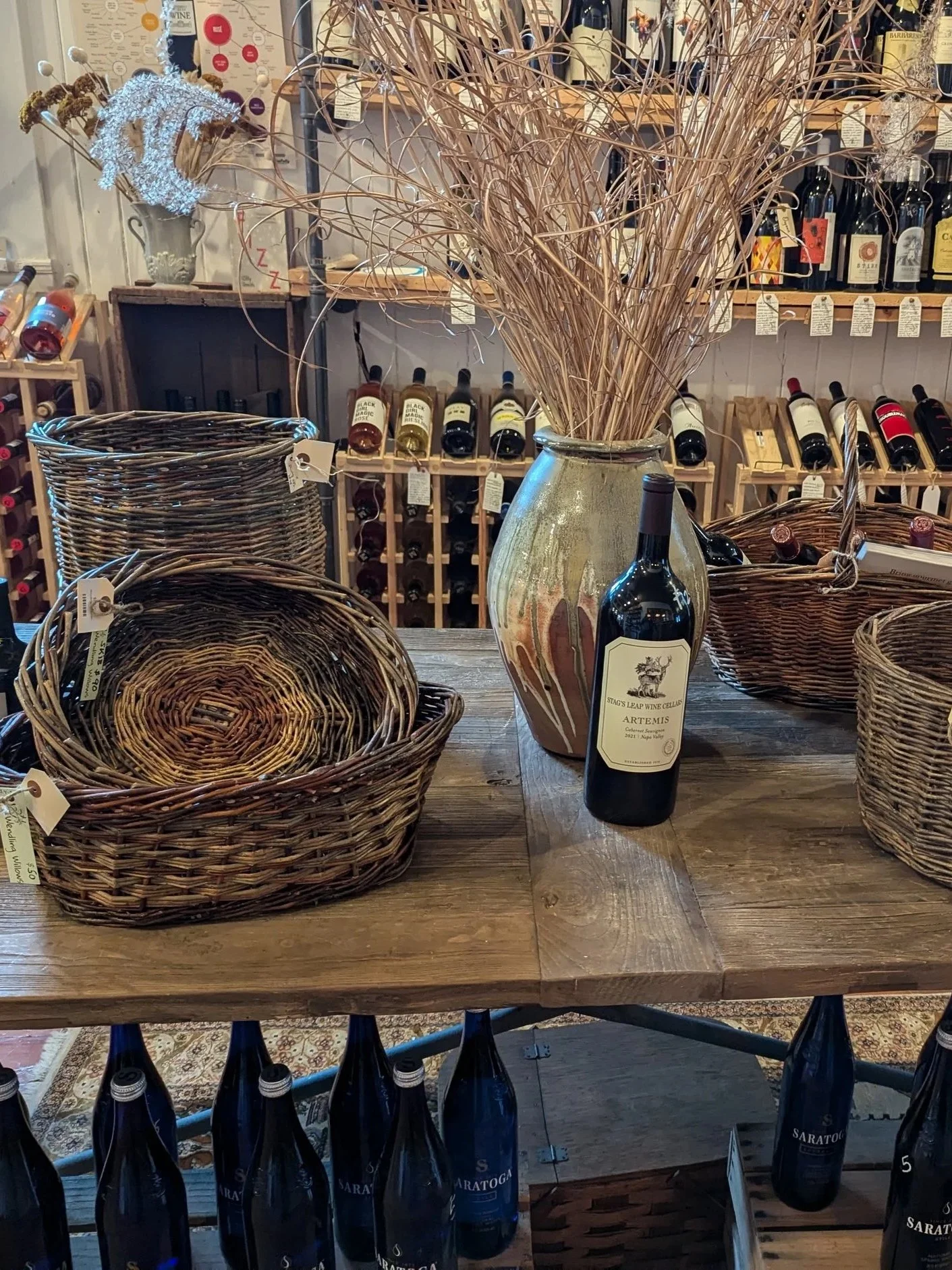 A rustic wooden table displaying a large ceramic vase filled with dried curly twigs, a bottle of Artemis wine, and several woven Wendling Willow baskets. 