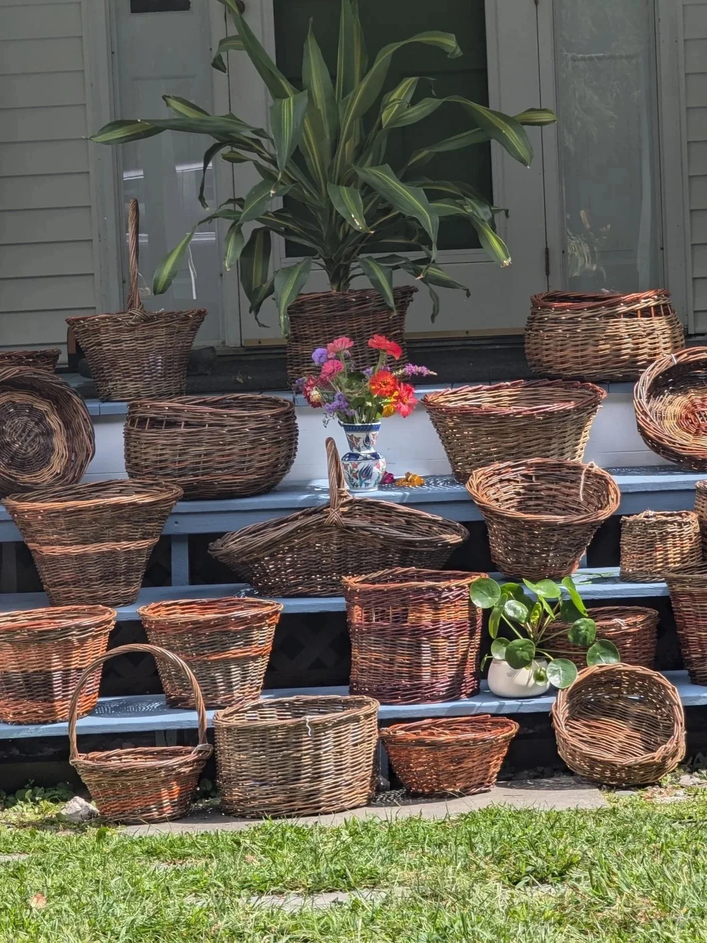 Various woven baskets arranged on steps outdoors, with a large green leafy plant, a vase of colorful flowers, and a small potted plant in the background.