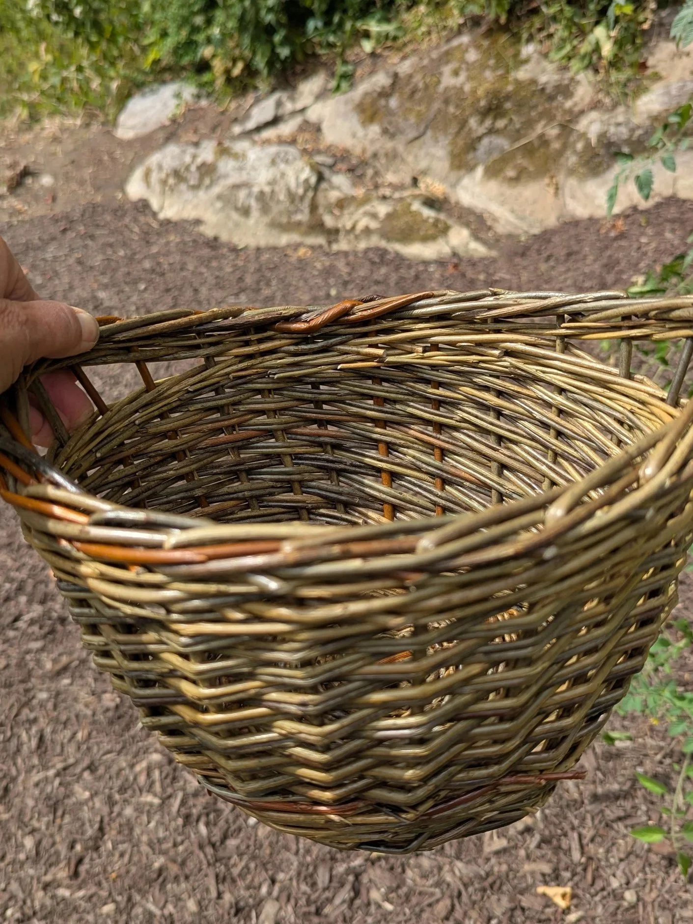 Close-up of a hand holding an empty woven basket outdoors with rocks and soil in the background.