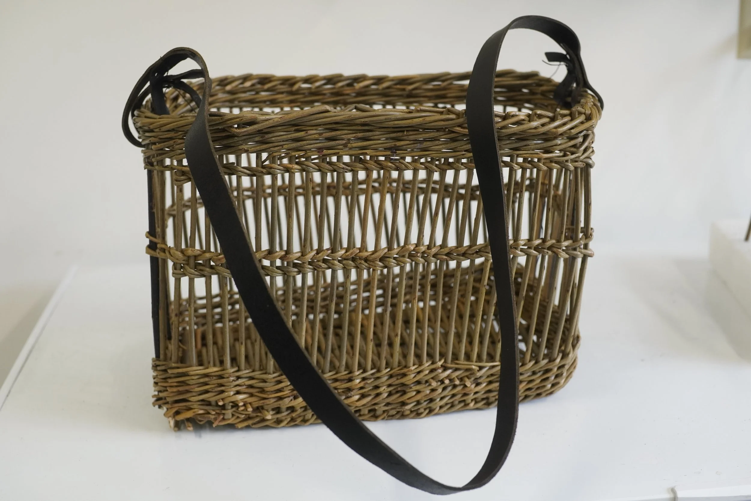 A woven wicker basket with black leather handles on a white surface against a white background.