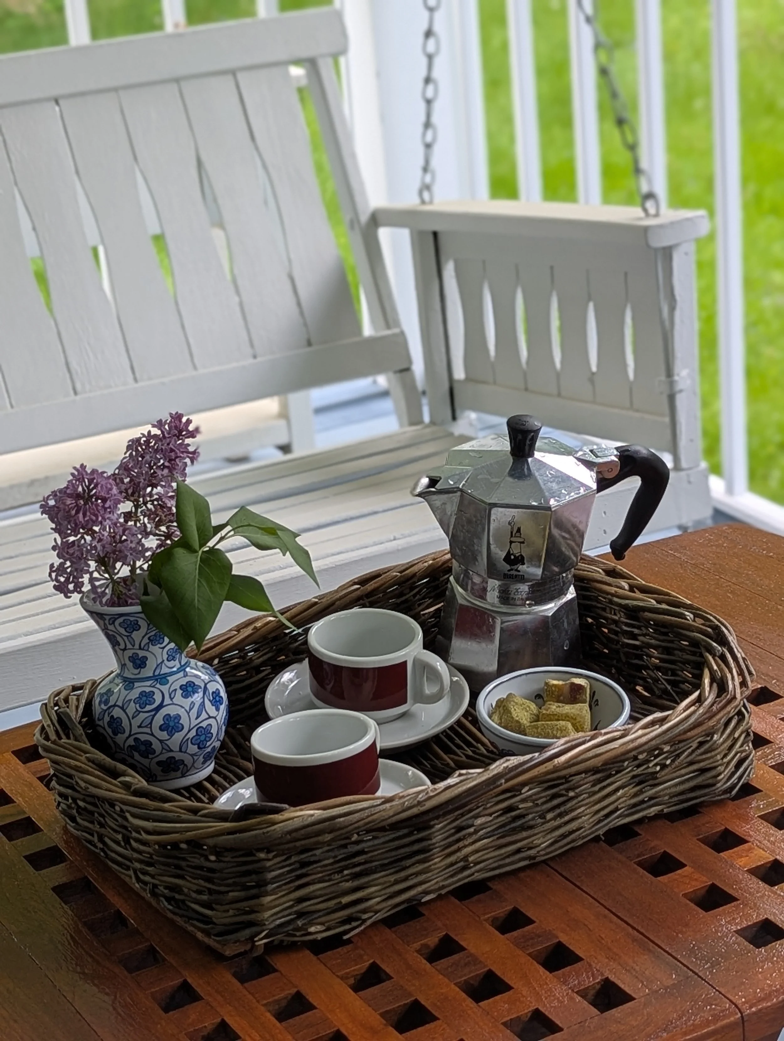A wicker tray on a wooden table holding a moka pot, two cups with saucers, a bowl of sugar cubes, and a small potted lilac plant, with a white porch swing and green grass in the background.