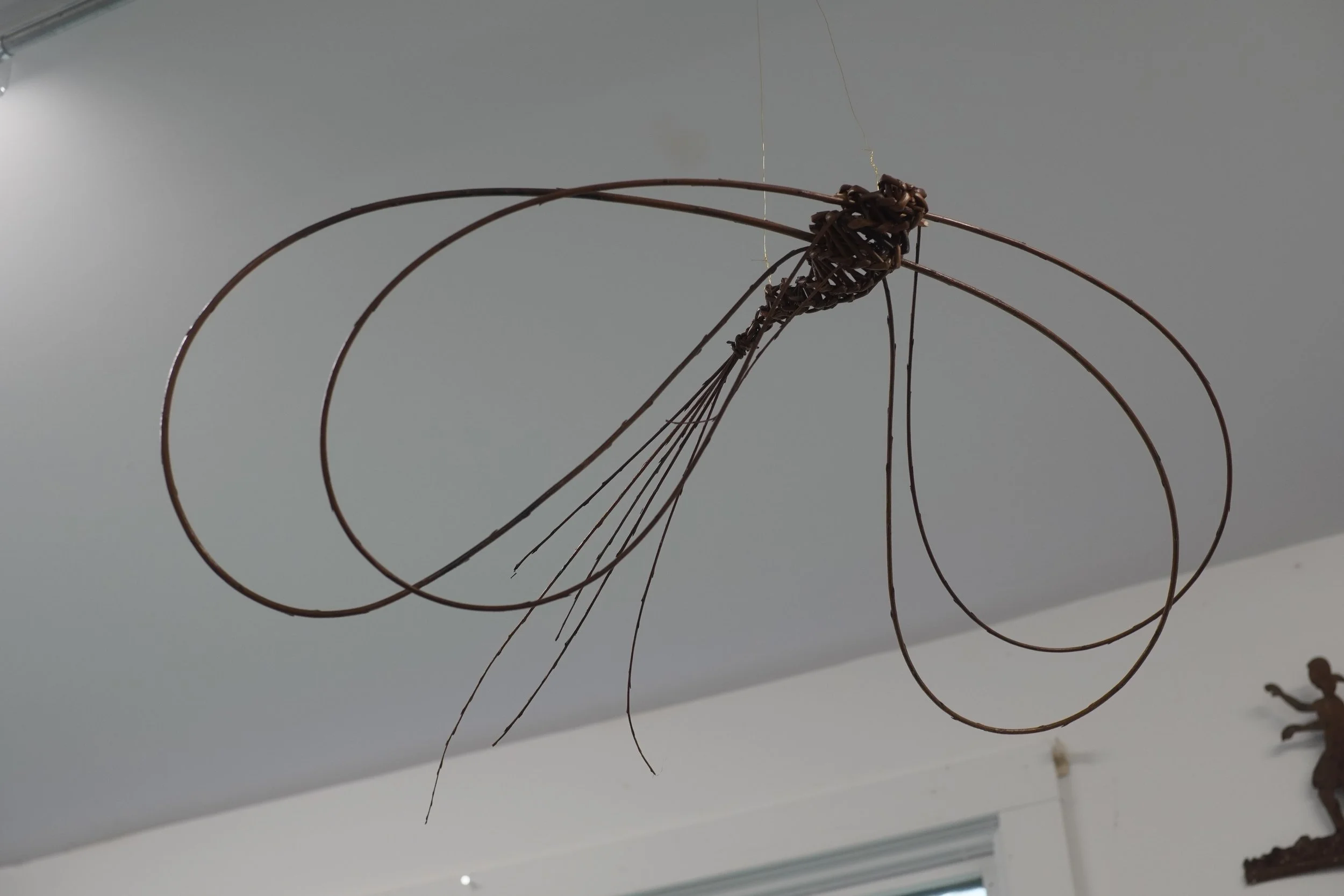 Metal sculpture of a hanging fish with elongated, curved fins and a textured body, displayed indoors on a white wall, with a small sculpture of a person pointing in the background.
