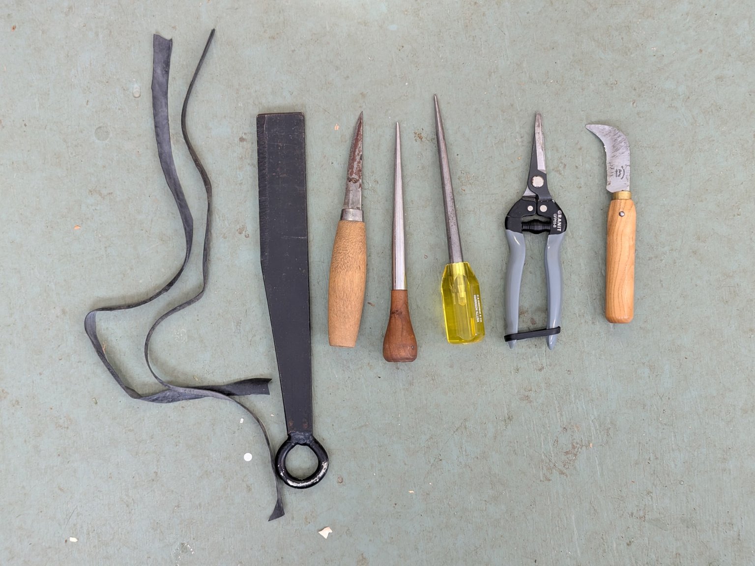 A collection of hand tools laid out on a green surface, including two wrenches, a large chisel, a small chisel, a screwdriver with a yellow handle, and a pruning shear with gray handles.