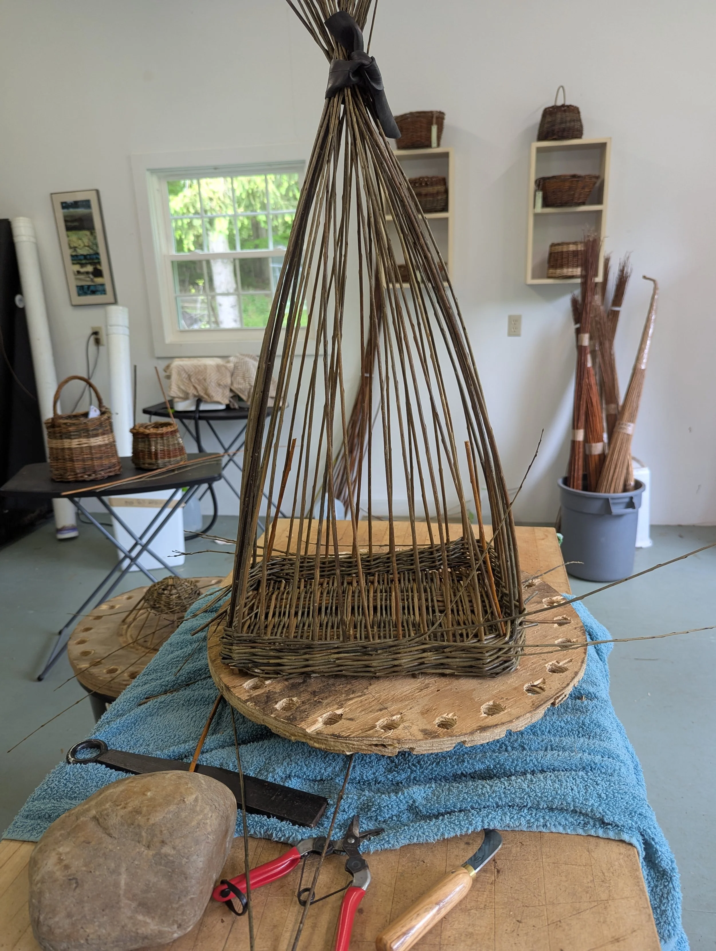 Wicker work in progress on a worktable in a craft room with tools, a rock, and other wicker pieces.