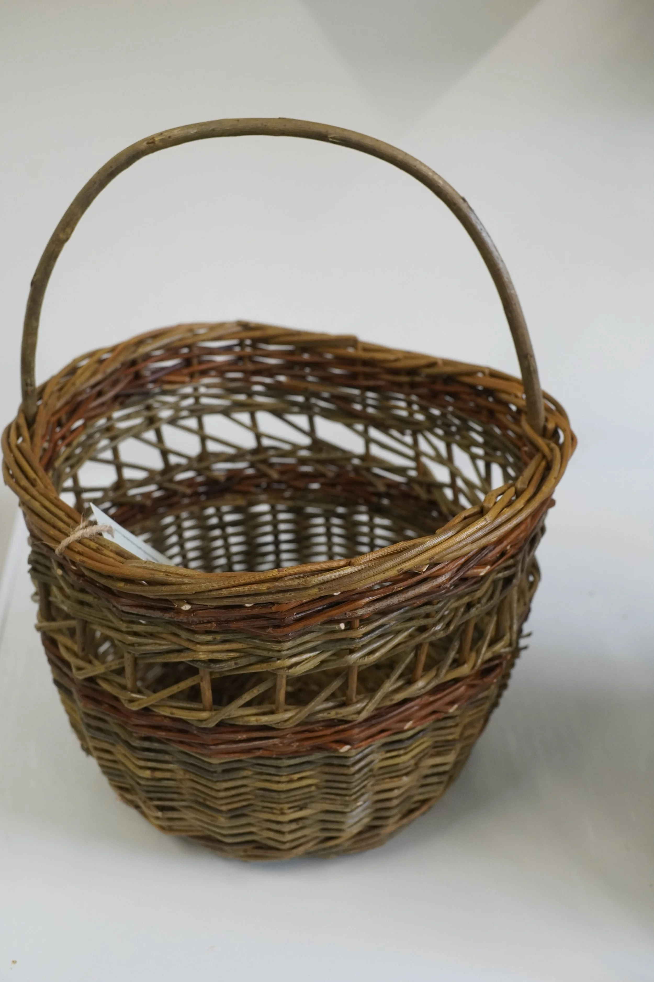 A woven wicker basket with a curved handle, placed on a white surface with a plain light grey background.