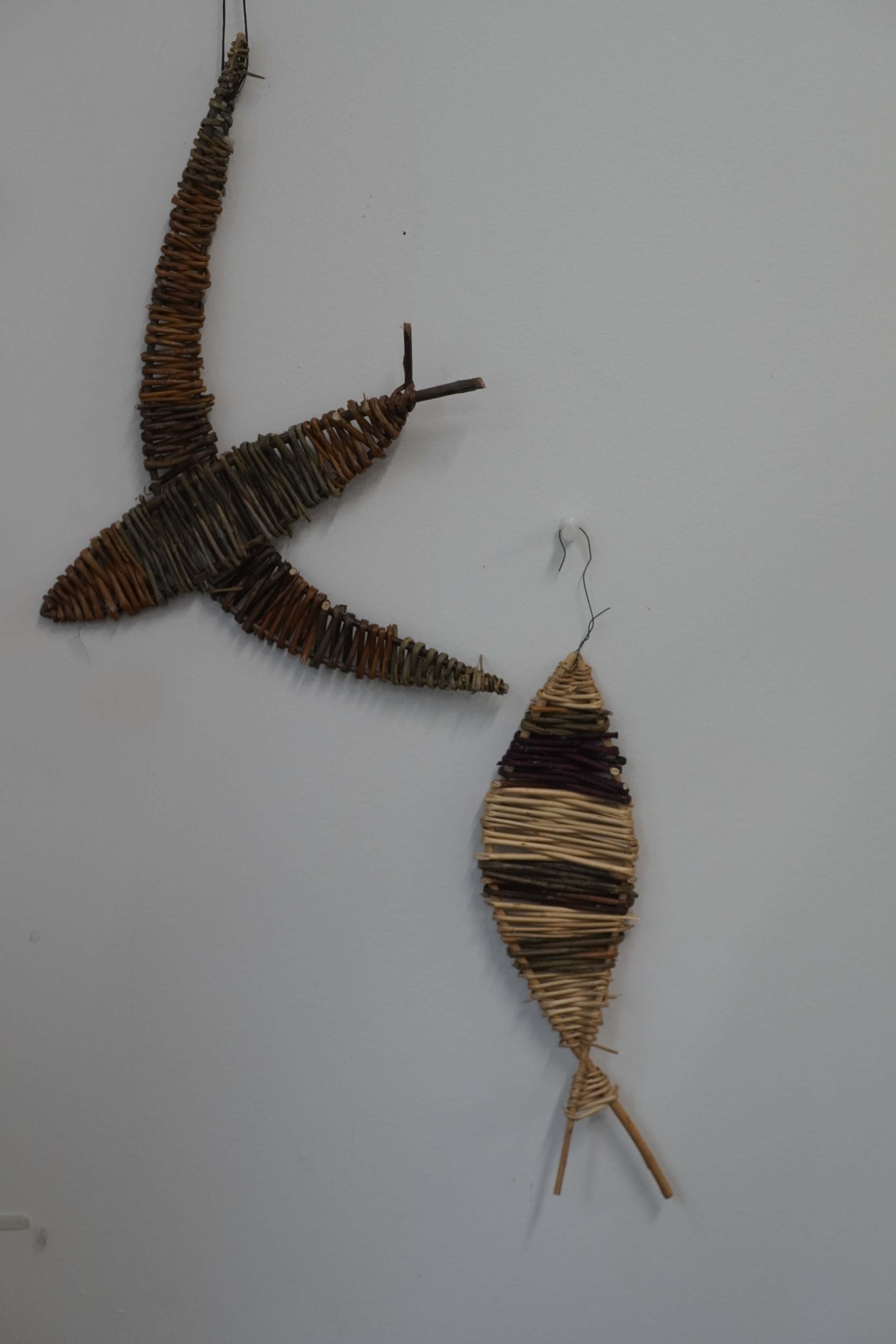 Wall art made of woven sticks resembling a fish or whale with a smaller woven piece hanging below.