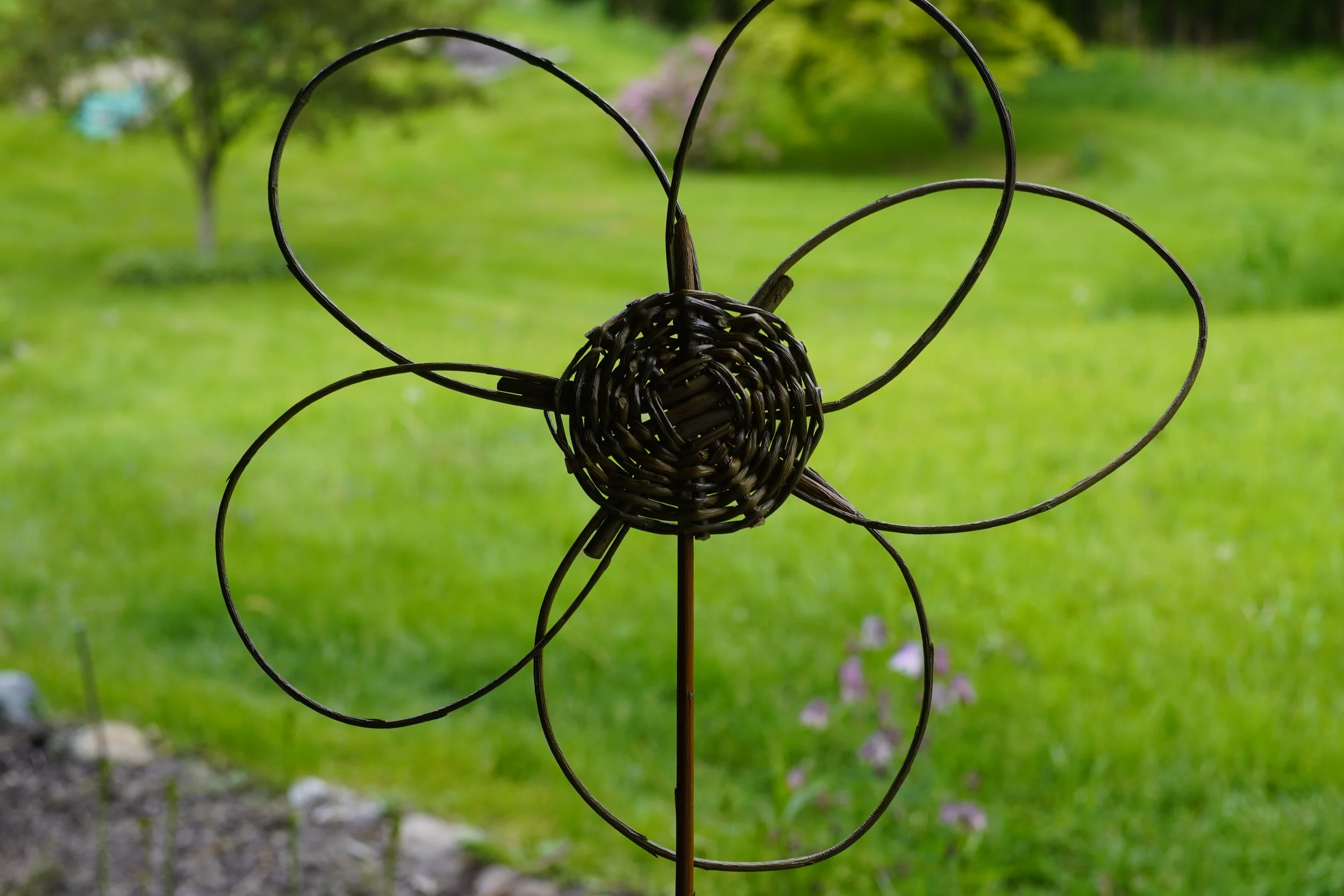Decorative garden wind spinner made of intertwined metal wires with a heart shape and circular design, set against a blurred green garden background.
