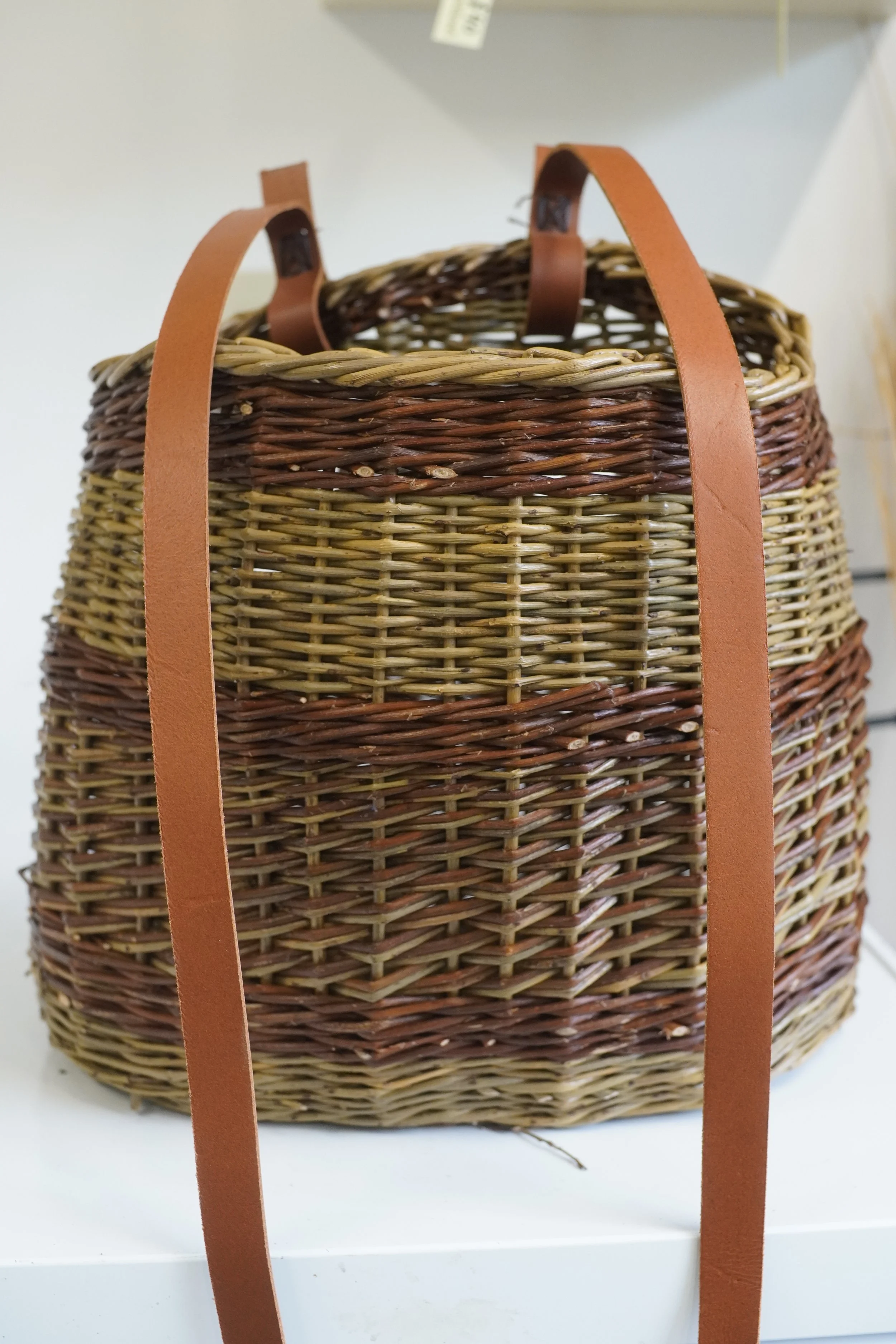 Woven wicker basket with leather straps on a white surface.