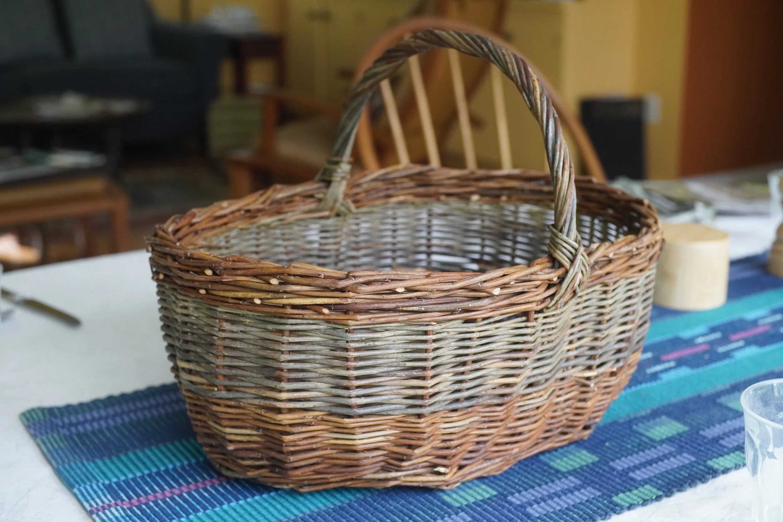 Stacked woven wicker baskets on a table, with a colorful striped tablecloth underneath. The background shows a room with chairs and shelves.