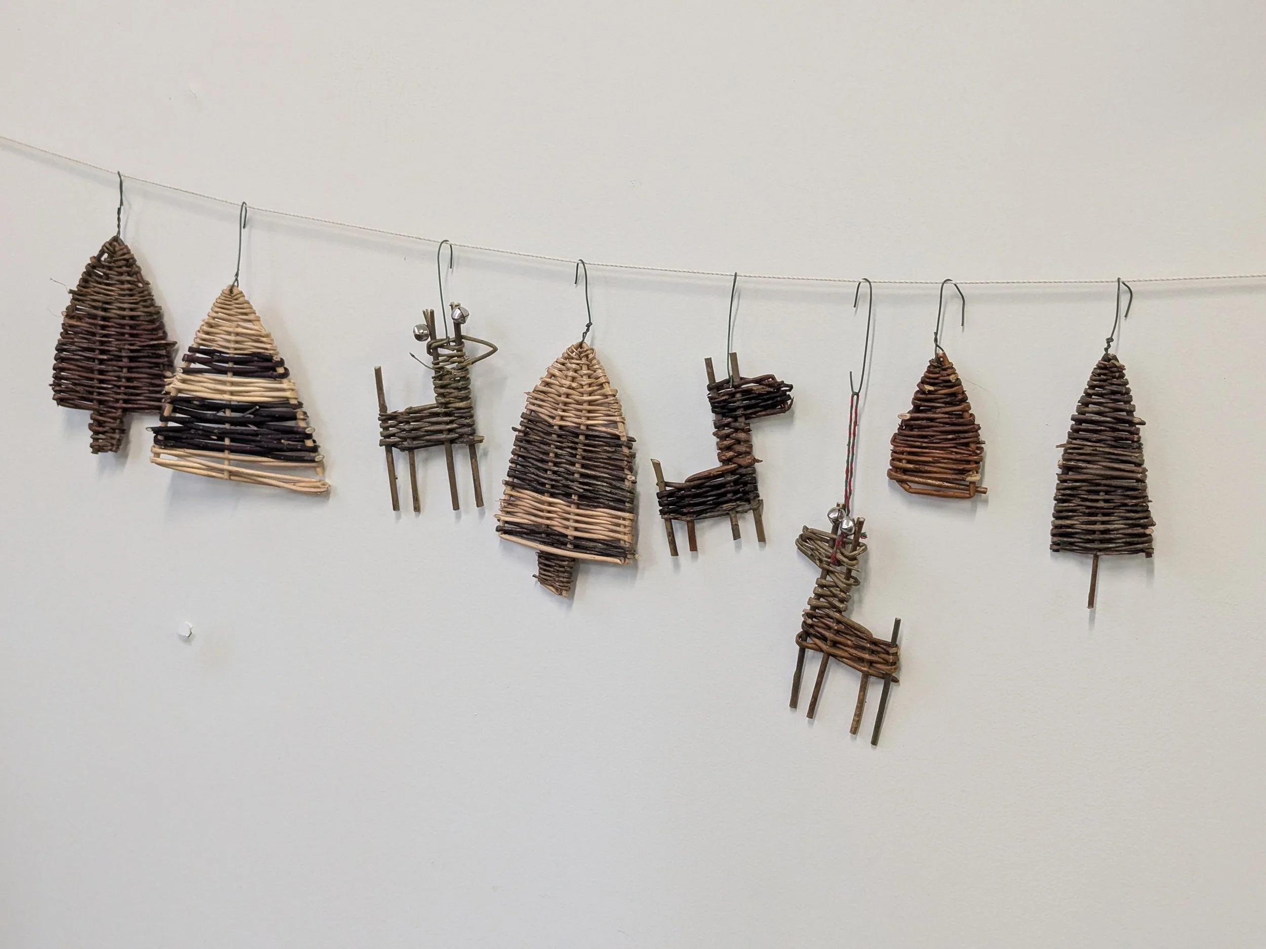 String of small woven wicker reindeer and Christmas trees hanging on a wire against plain white wall.