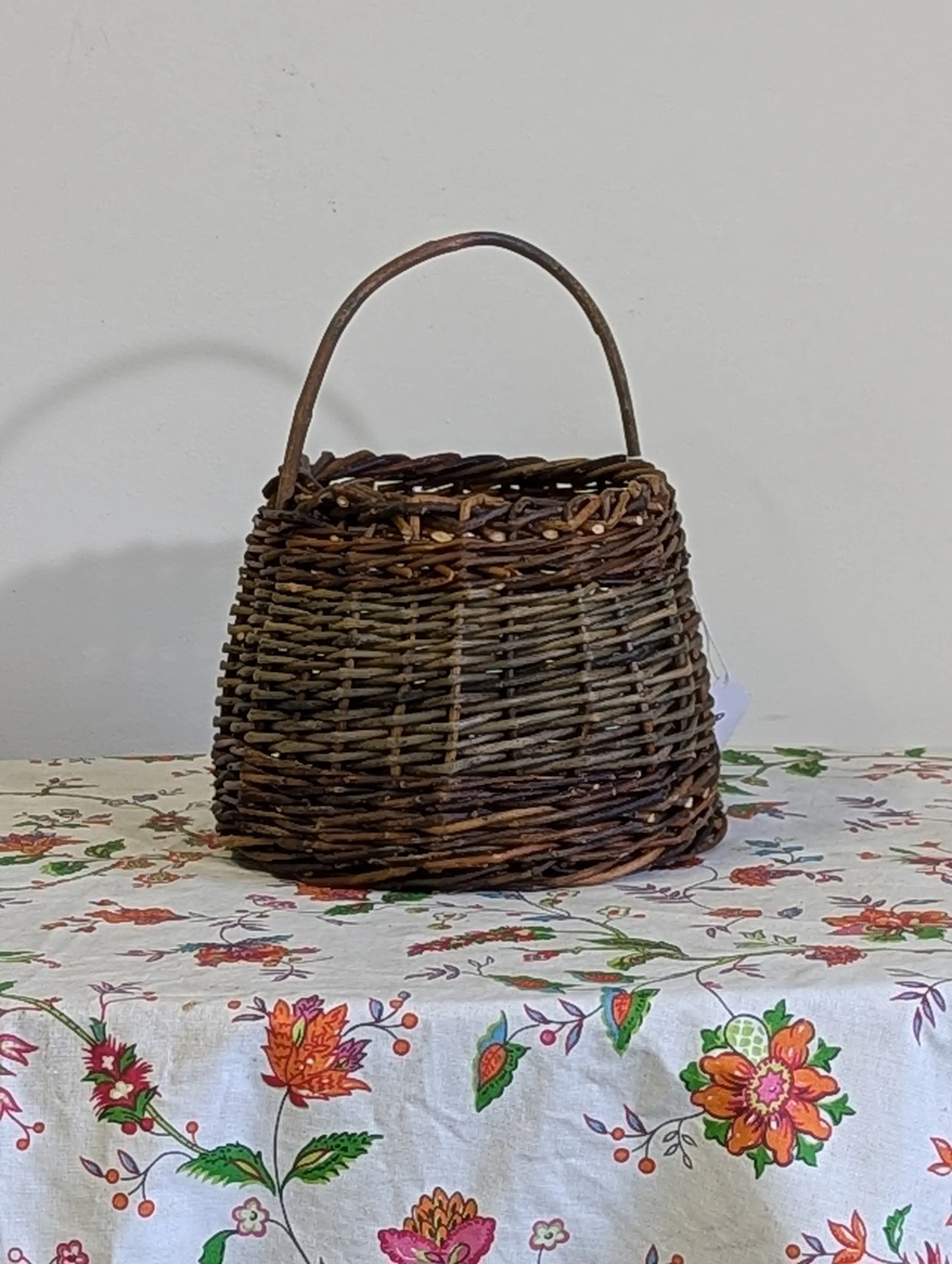 A woven wicker basket sitting on a table with a floral tablecloth, against a plain white wall background.