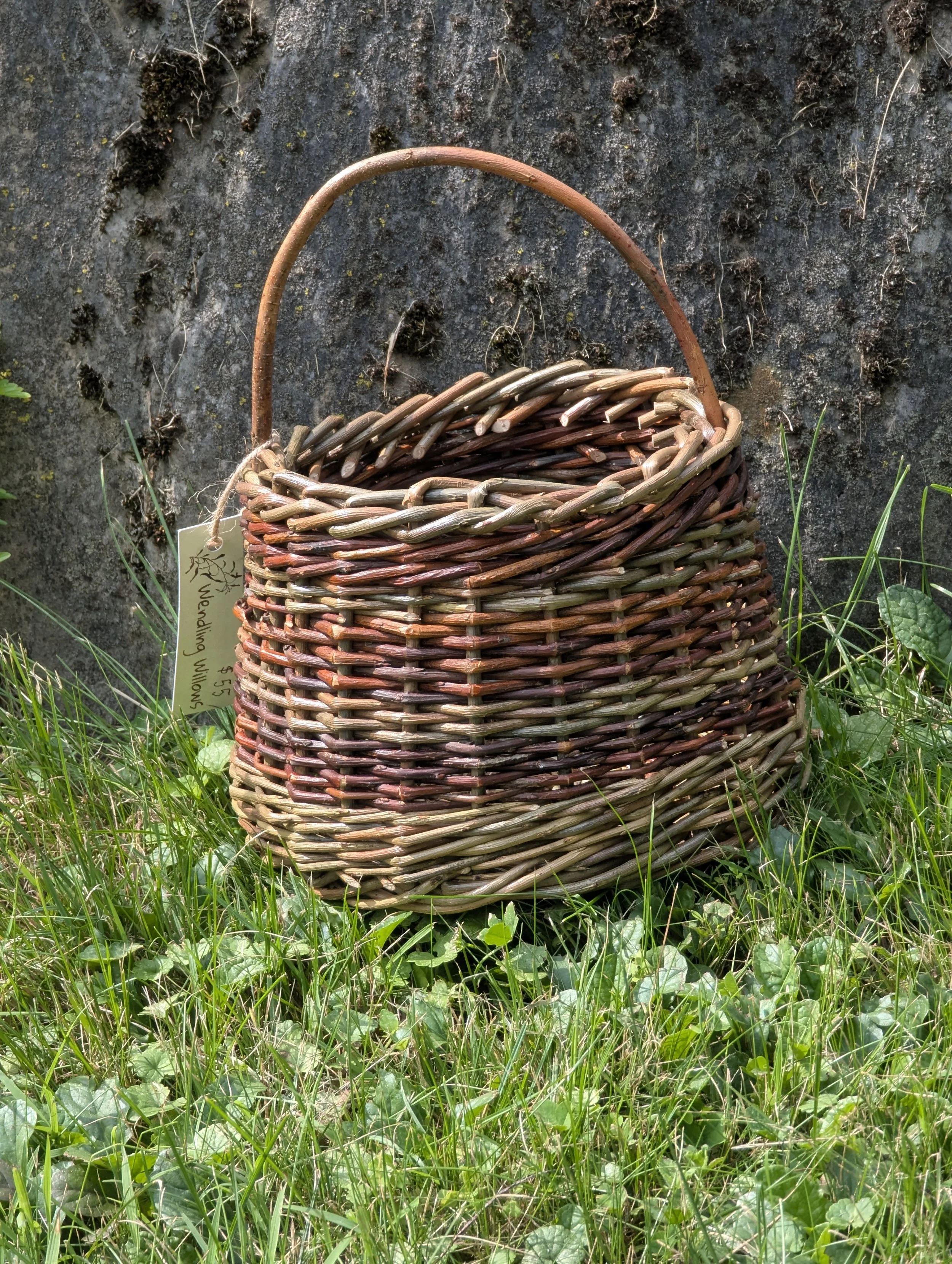 Handwoven willow basket sitting on grass near a stone wall, with a tag attached to the basket that says 'Wendling Wows'.