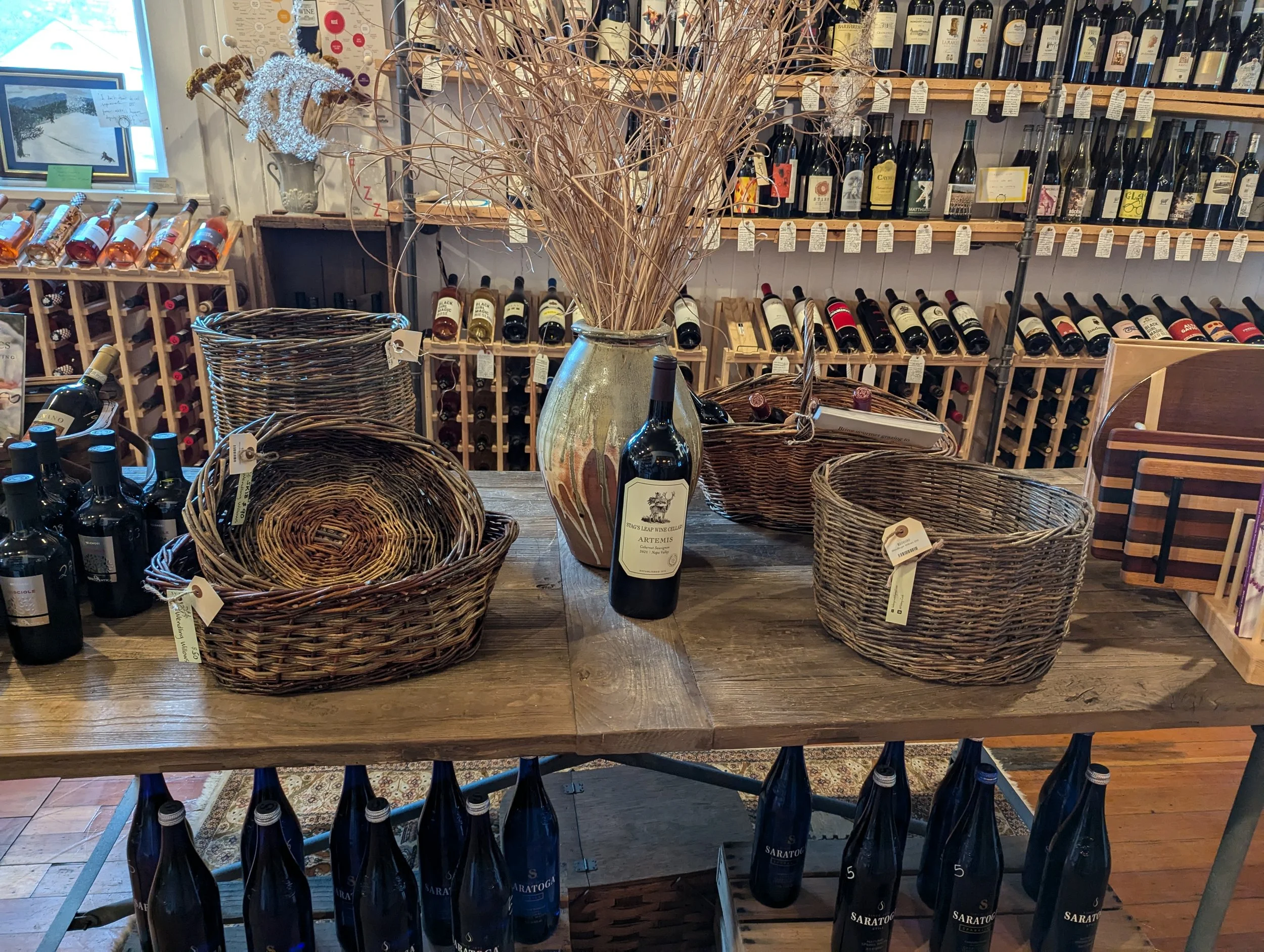 A rustic wooden table displays empty woven baskets, a large ceramic vase with dried grasses, and a bottle of wine labeled 'Artemis' in a wine shop. In the background, shelves are stocked with various bottles of wine.