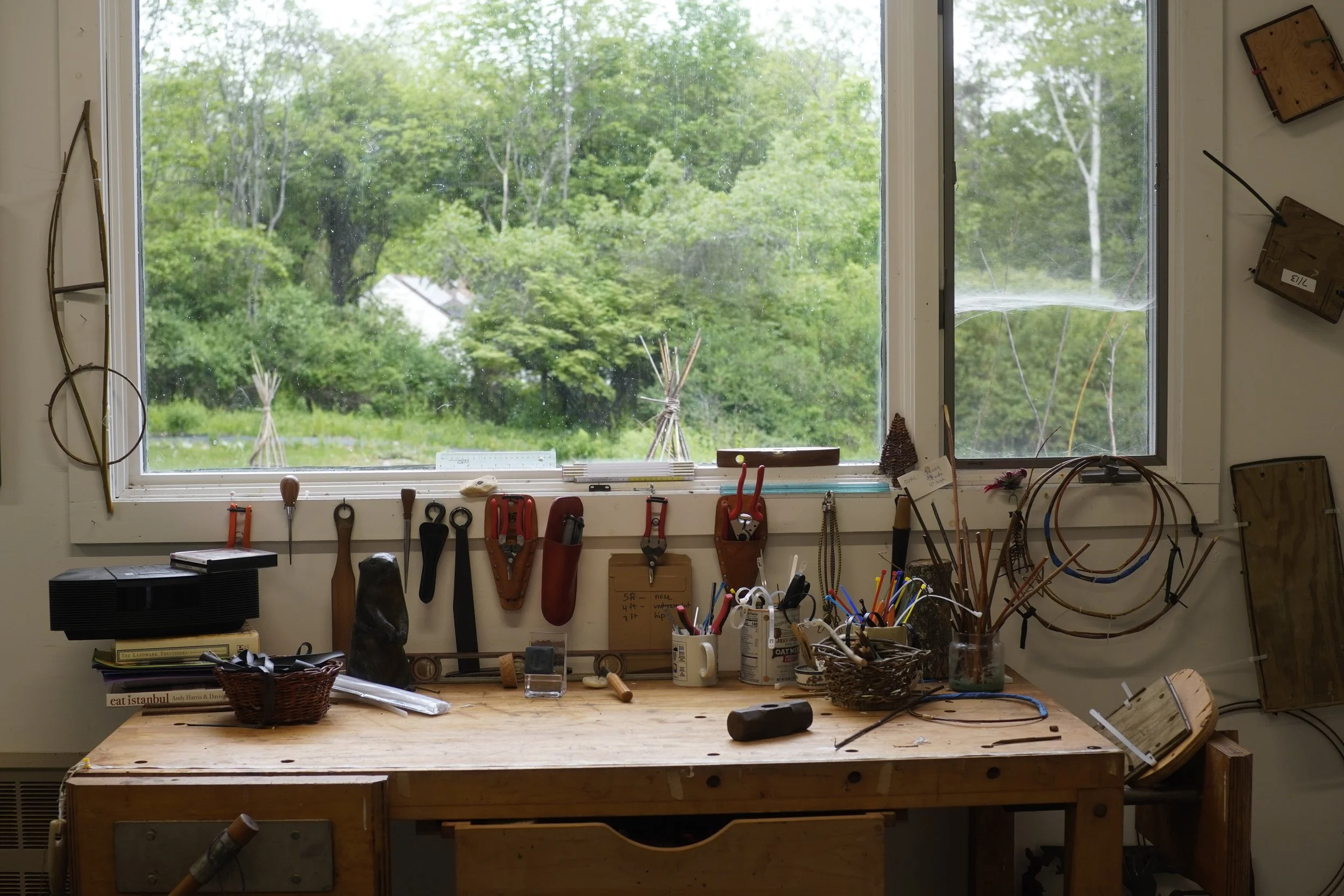 View of a cluttered workbench with various tools, baskets, and jars in a workshop, overlooking a window with a lush green outdoor scene.
