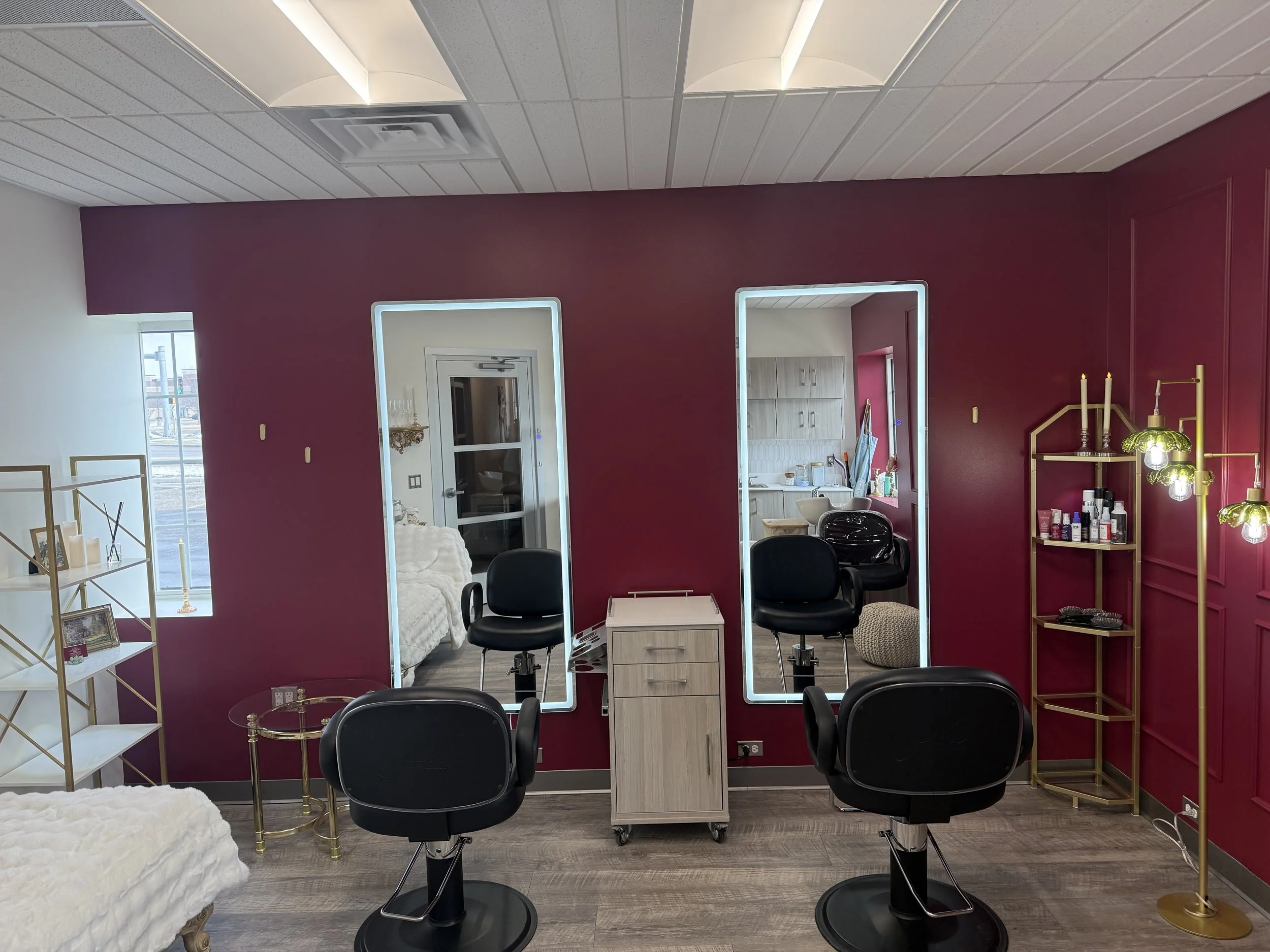 The Hair Wardrobe & Parlor, Janesville WI.  Salon with two black styling chairs in front of two large illuminated mirrors. To the right is a gold shelving unit with beauty products, and a floor lamp with multiple light bulbs.