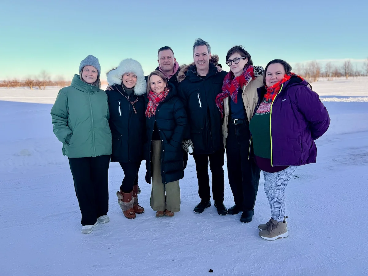 Seven people stand together for a photo on snow-covered ground, several wearing traditional Sámi scarves