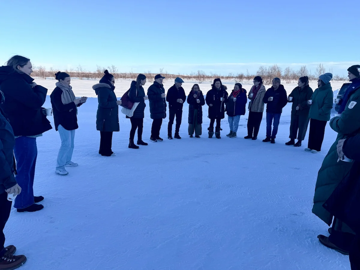 A large group stands in a wide circle on a snow-covered plain under a clear blue sky, holding cups
