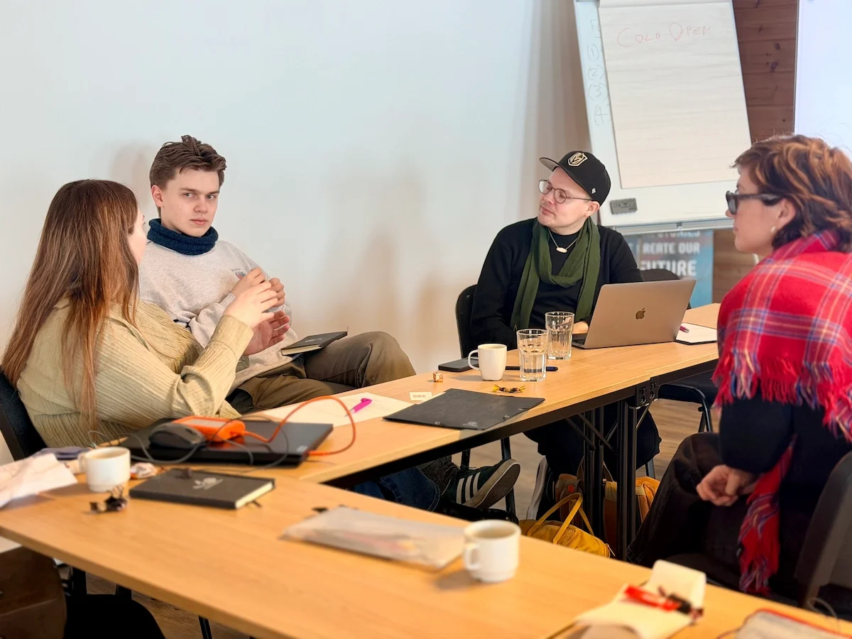 Four workshop participants in close conversation around a table, one taking notes on a laptop
