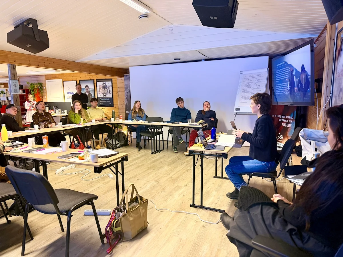 A workshop leader speaks to participants seated around tables, beside a banner reading "By telling our stories we create our future"